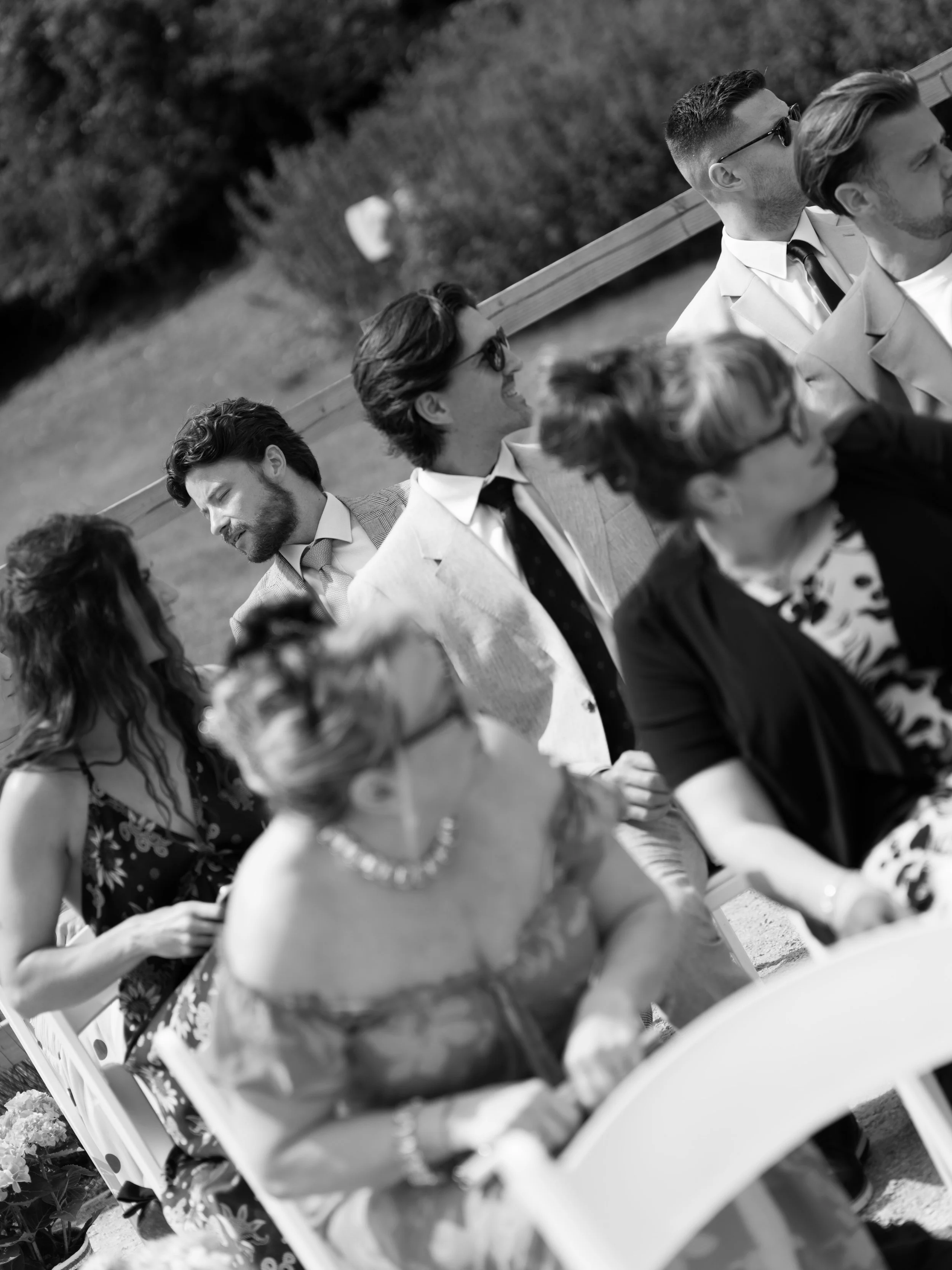 Black and white photo of four people sitting on a bench outdoors. They are dressed formally, some wearing glasses and ties, and are smiling or looking away. There is greenery in the background.