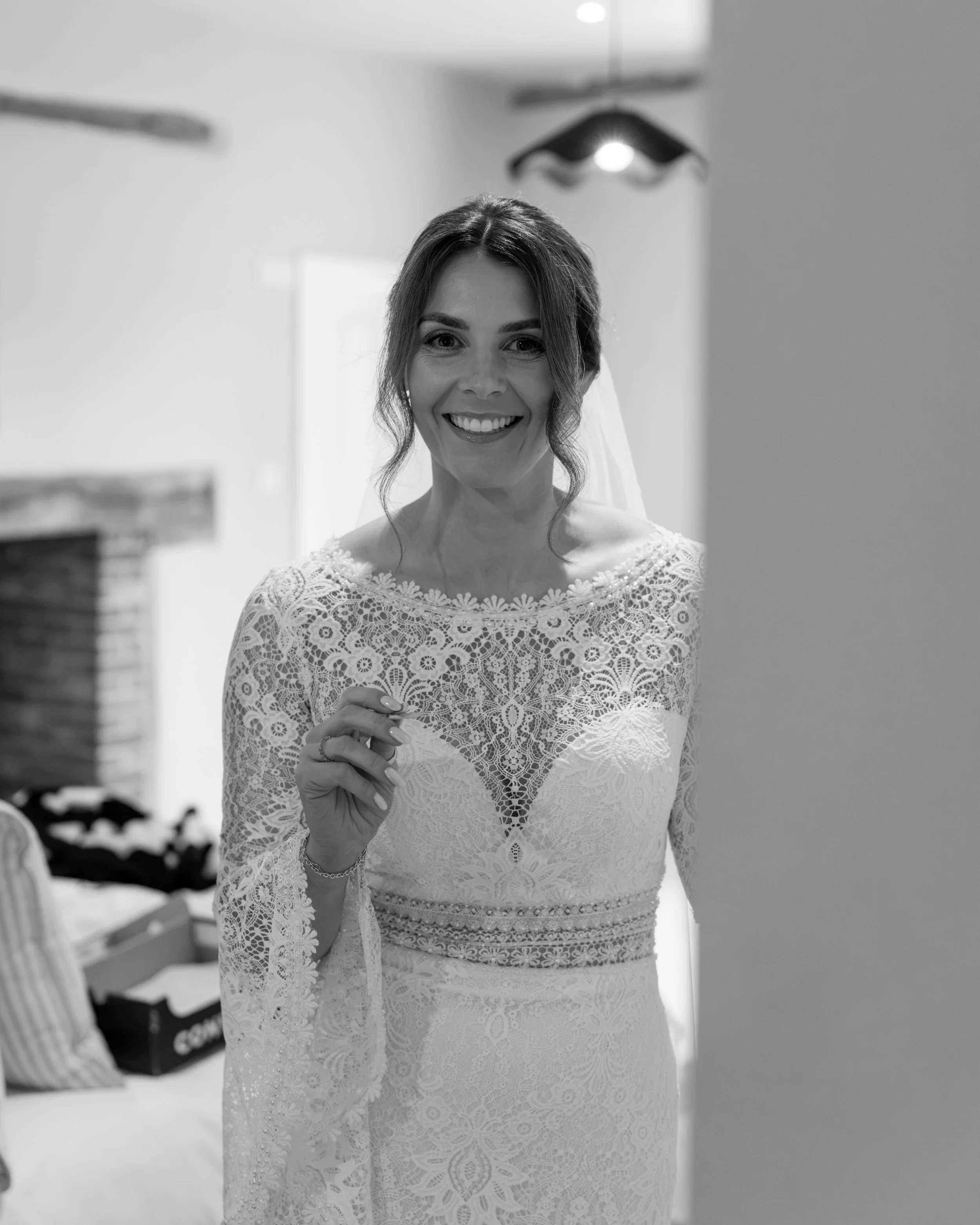 Black and white photo of a woman wearing a lace dress, smiling, standing indoors with light coming from above, background includes a wall and some stacked objects.