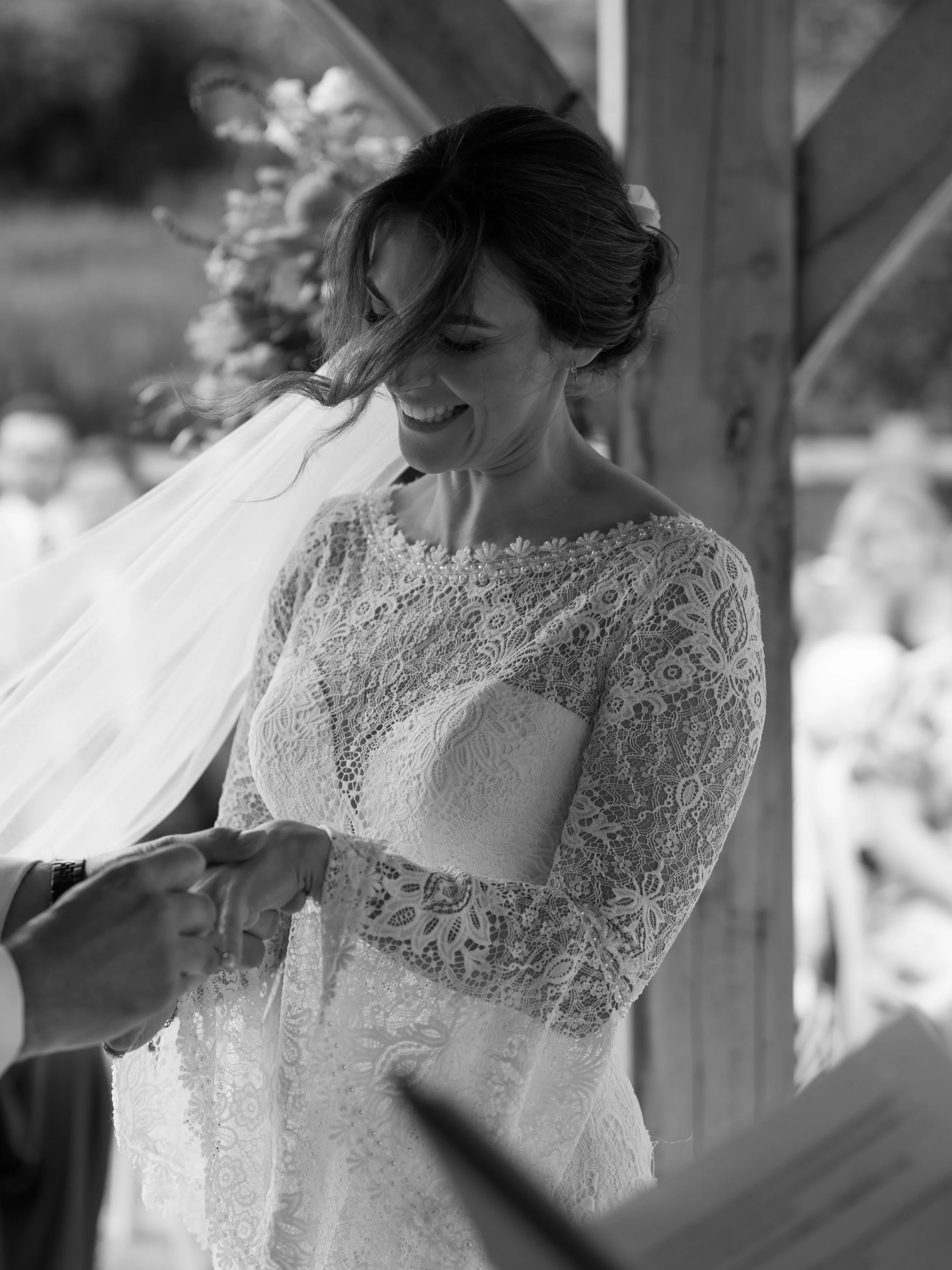 Black and white photo of a bride smiling as someone adjusts her wedding dress and veil.