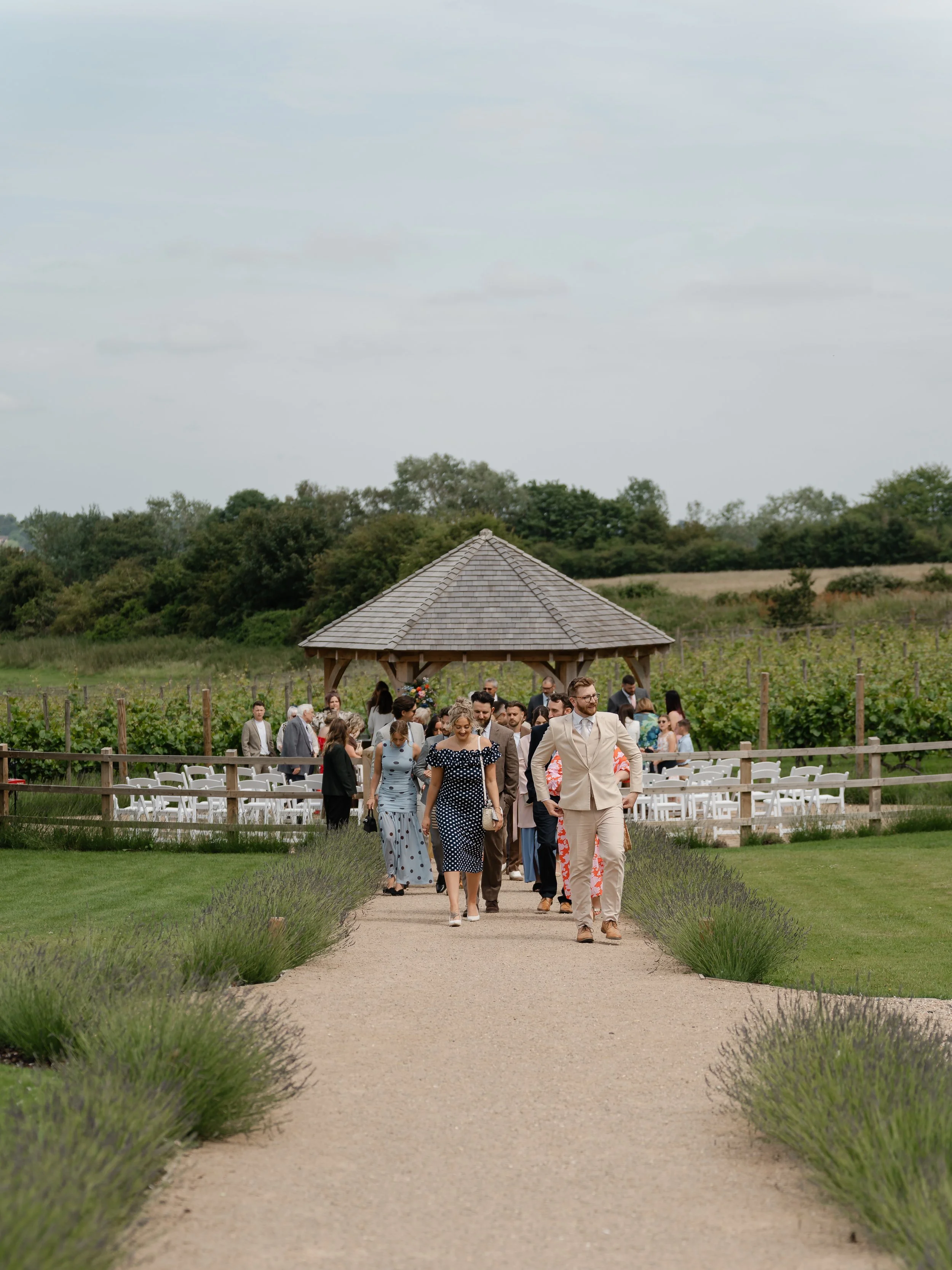 People dressed in formal attire walking along a vineyard path towards the outdoor wedding ceremony area at Copdock Hall with white chairs, wooden fence, and a thatched-roof pavilion in a scenic countryside setting.