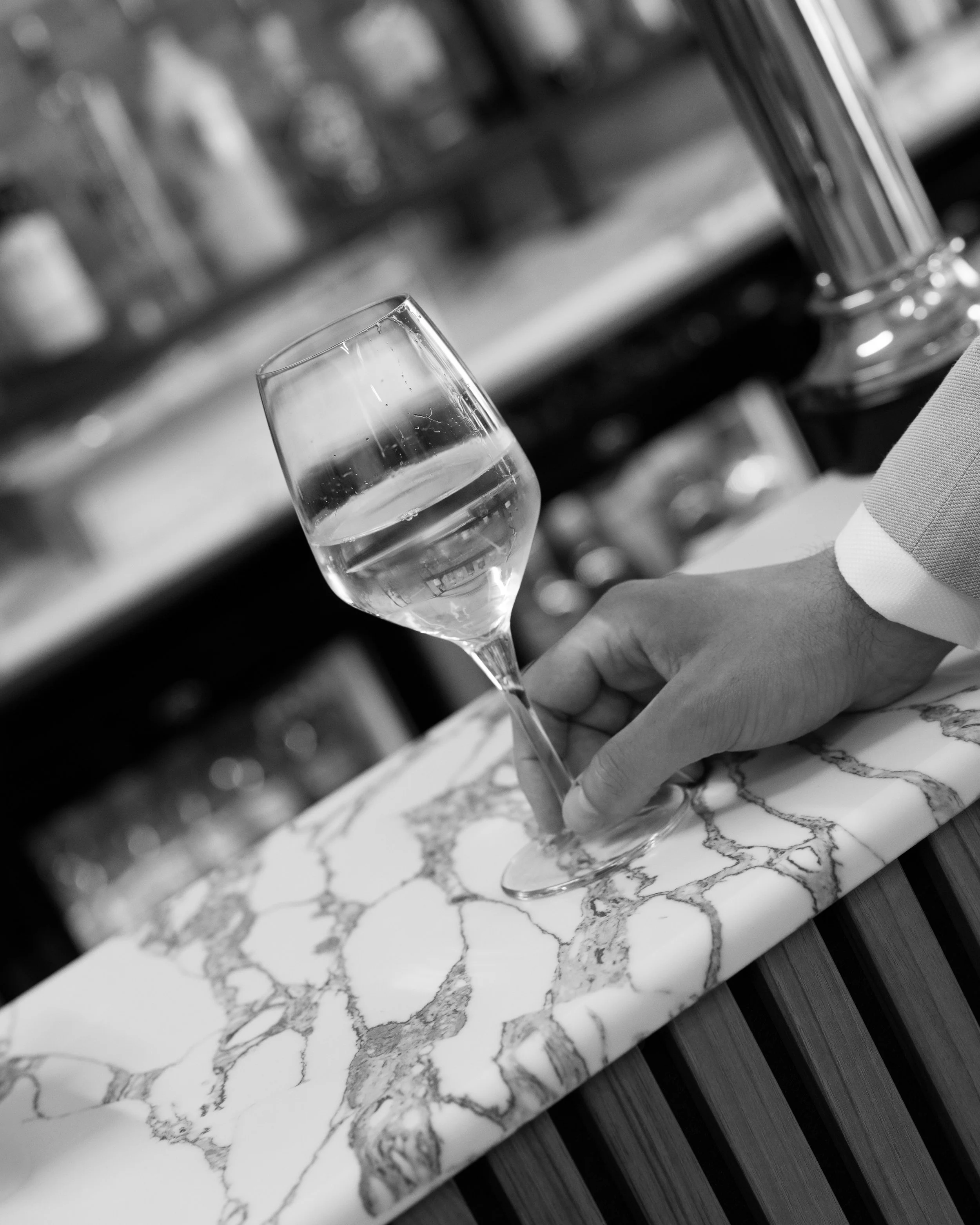 A person holding a glass of white wine or champagne over a marble countertop, with a blurred background of bottles on a shelf.