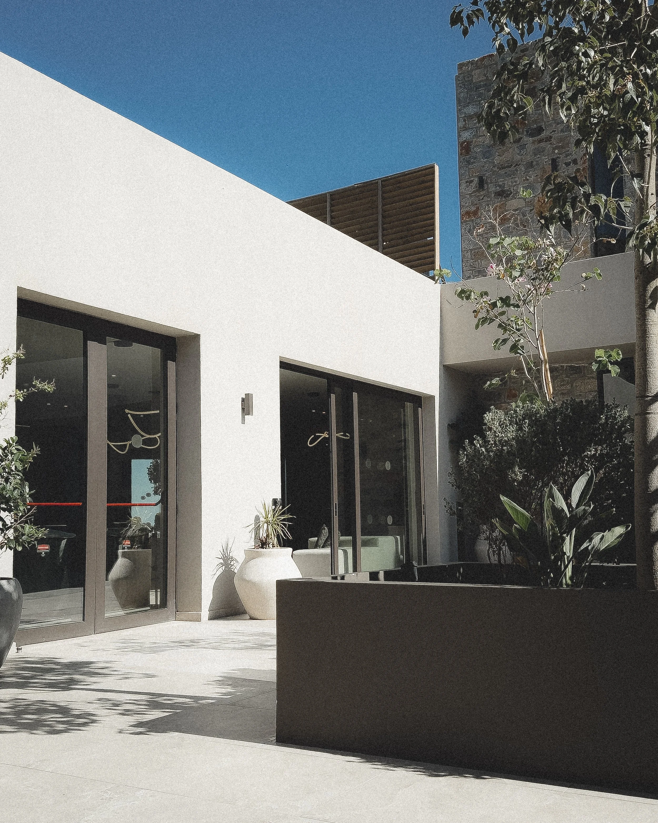 Exterior view of a modern house with white walls, large glass doors, and decorative planters, with a blue sky and trees in the background.