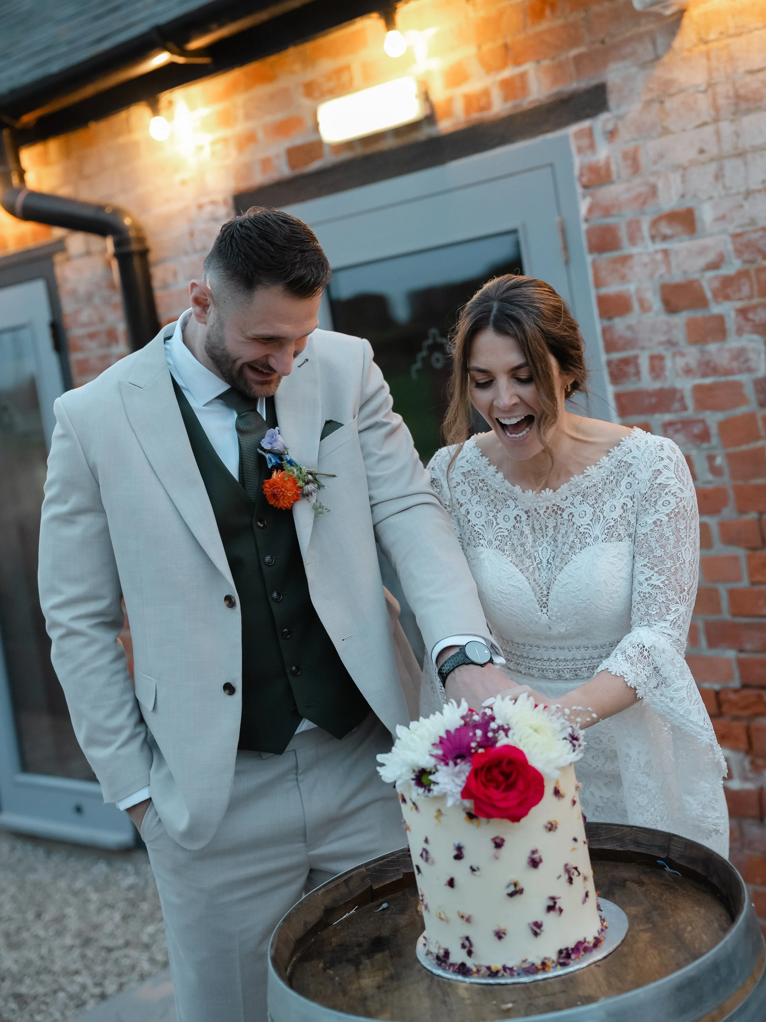 A bride and groom smiling and looking at a cake with flowers on top, during a wedding celebration outside near a brick building.