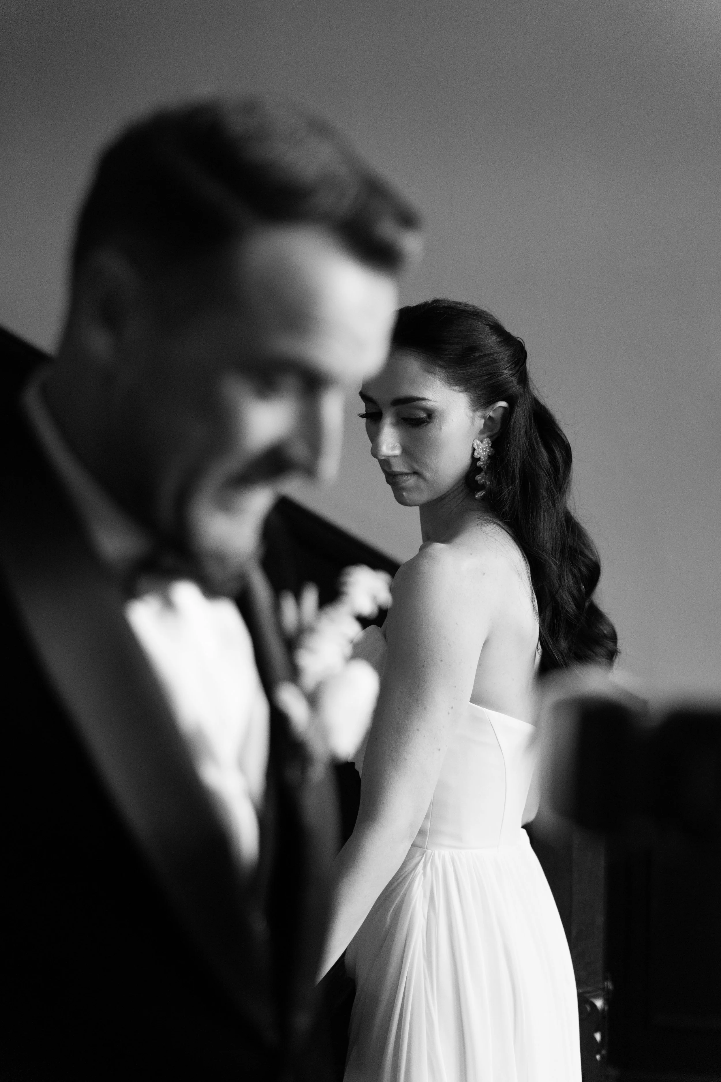 A black-and-white photo of a bride with long dark hair and earrings, looking down, standing behind a groom who is smiling and looking down, both dressed in wedding attire.