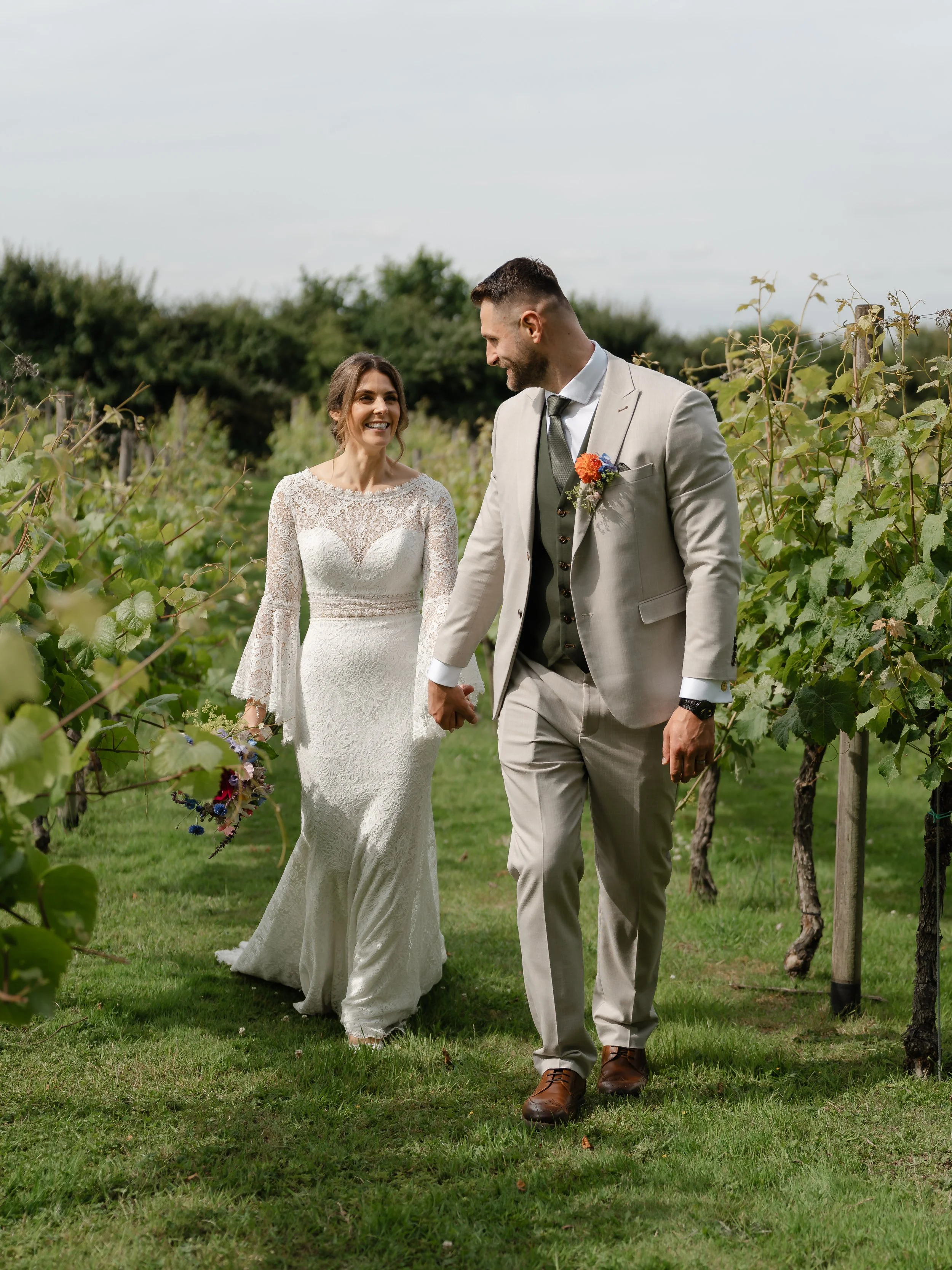 A bride and groom holding hands and smiling on their wedding day in an outdoor vineyard setting, with green foliage and grapevines around them.