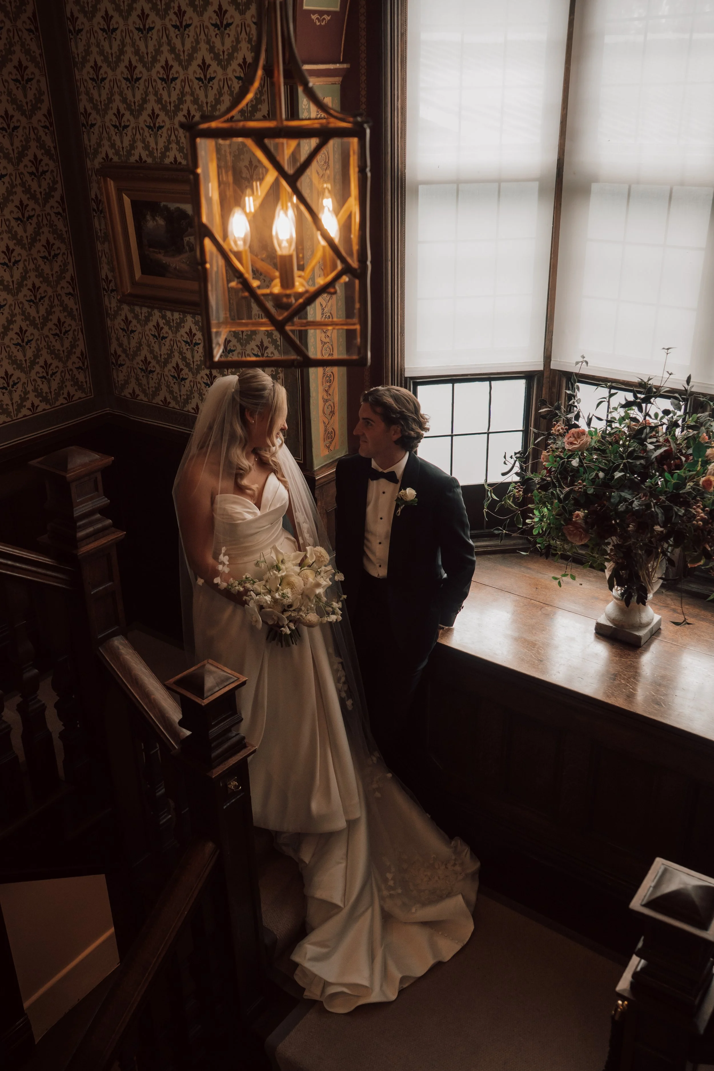 A bride and groom standing together inside a dimly lit, vintage-style room with dark wood paneling, floral wallpaper, and large windows. The bride is holding a bouquet of white flowers, and the groom is dressed in a tuxedo with a white boutonniere. T