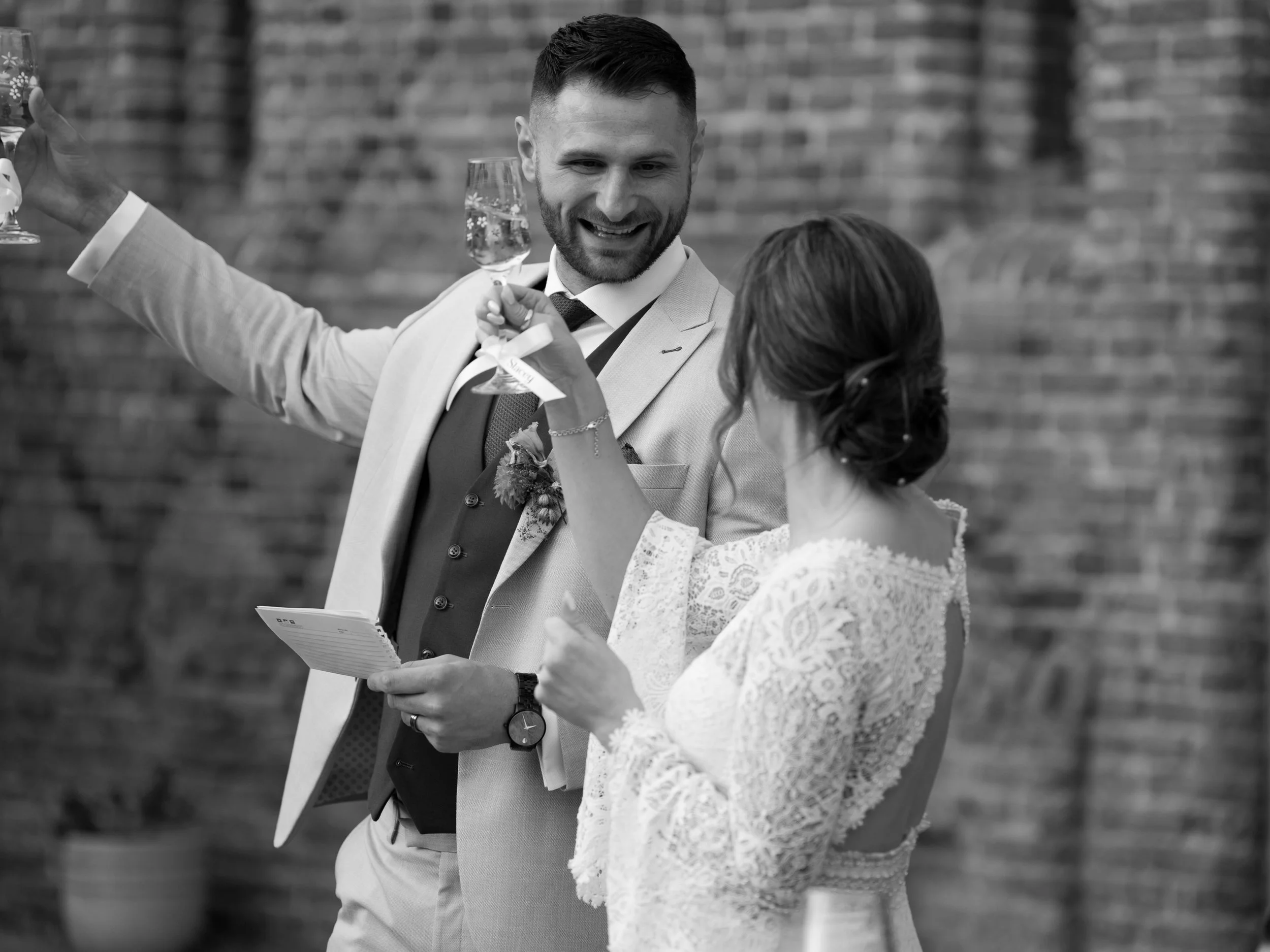 A smiling man in a light-colored suit and a woman in a lace dress holding glasses of champagne at a celebration or wedding.