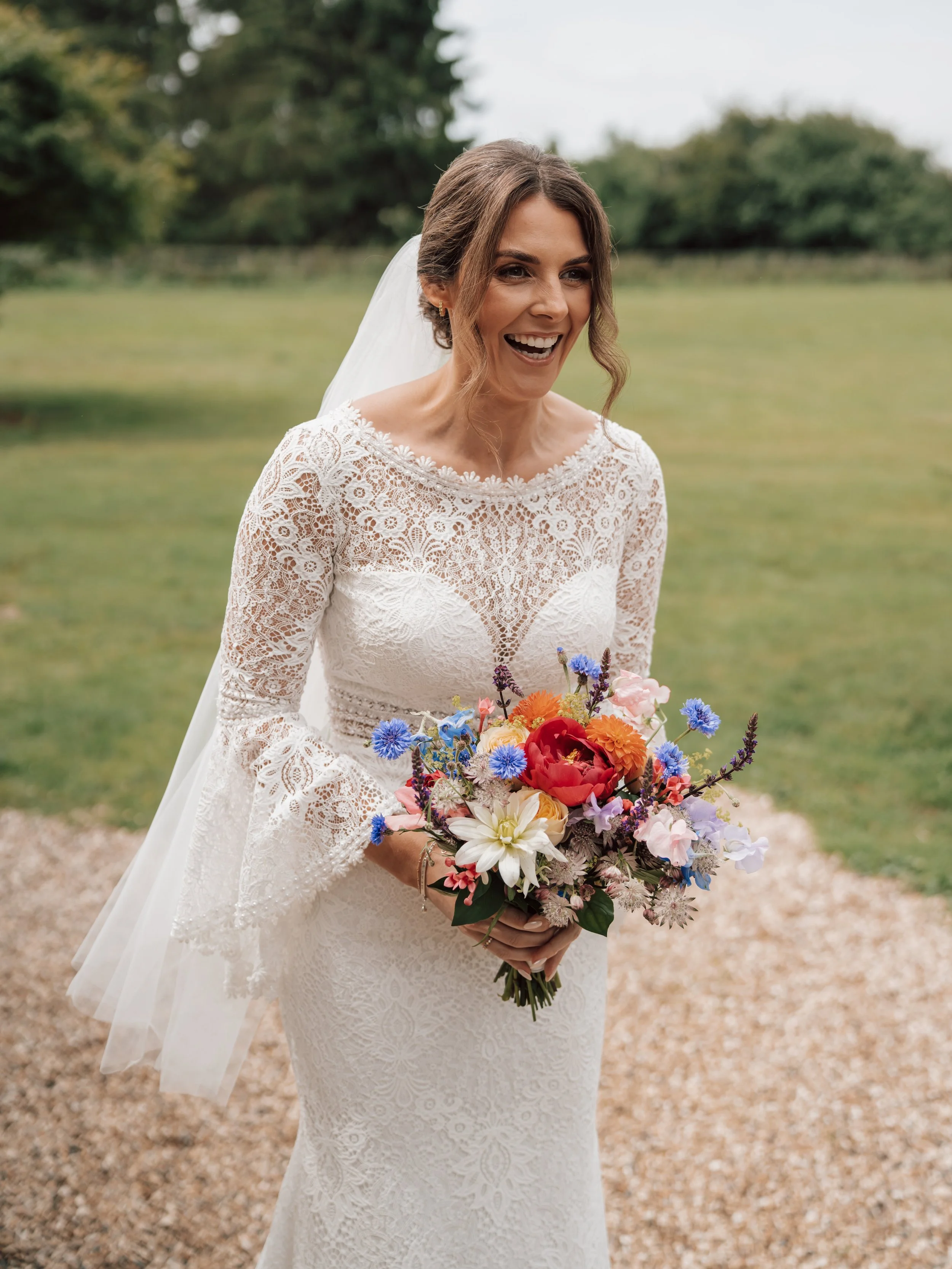 A bride in a white lace wedding dress holding a colorful bouquet of flowers, outdoors with greenery in the background.