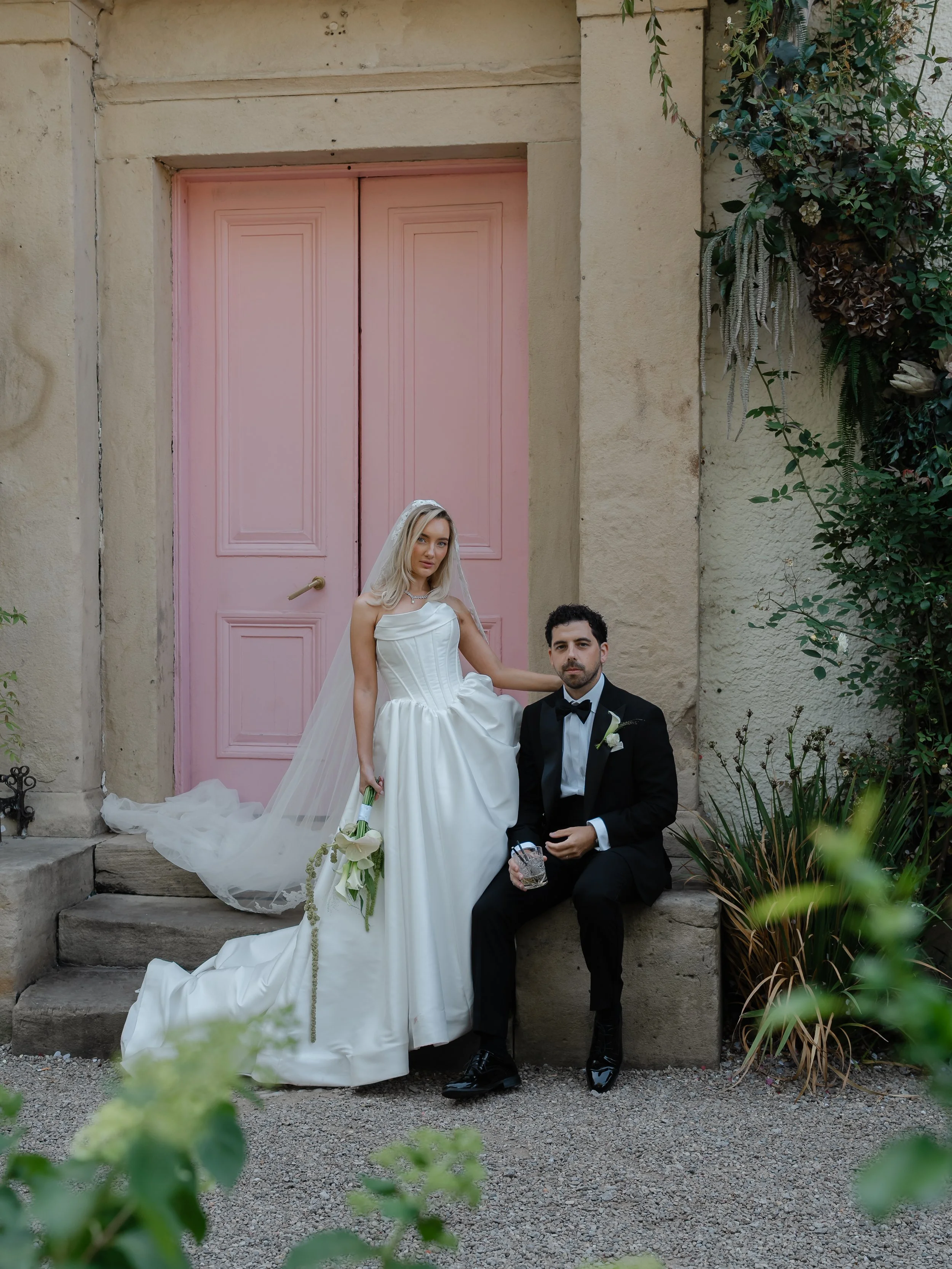 A bride in a white wedding dress and veil stands next to a groom in a black tuxedo, sitting on steps outside a building with a pink door and beige stone walls. The bride holds a bouquet of white flowers. They are surrounded by green plants.