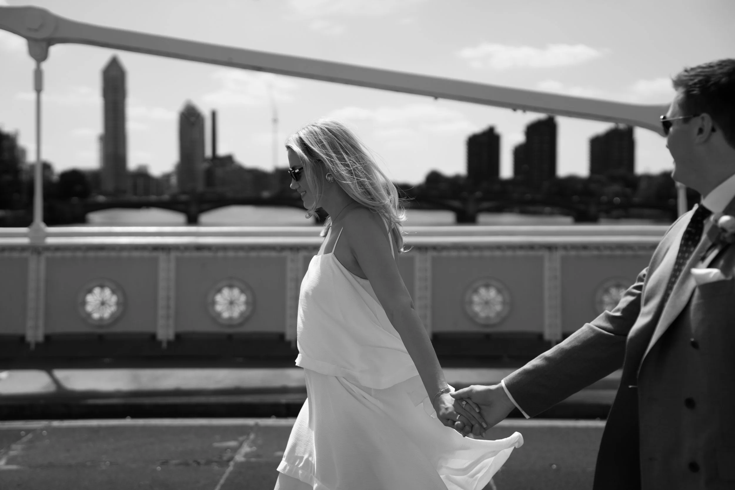 A black and white photo of a couple holding hands while walking across a bridge in an urban setting. The woman is wearing sunglasses and a white dress, and the man is wearing glasses and a suit. There are tall buildings and a city skyline in the back