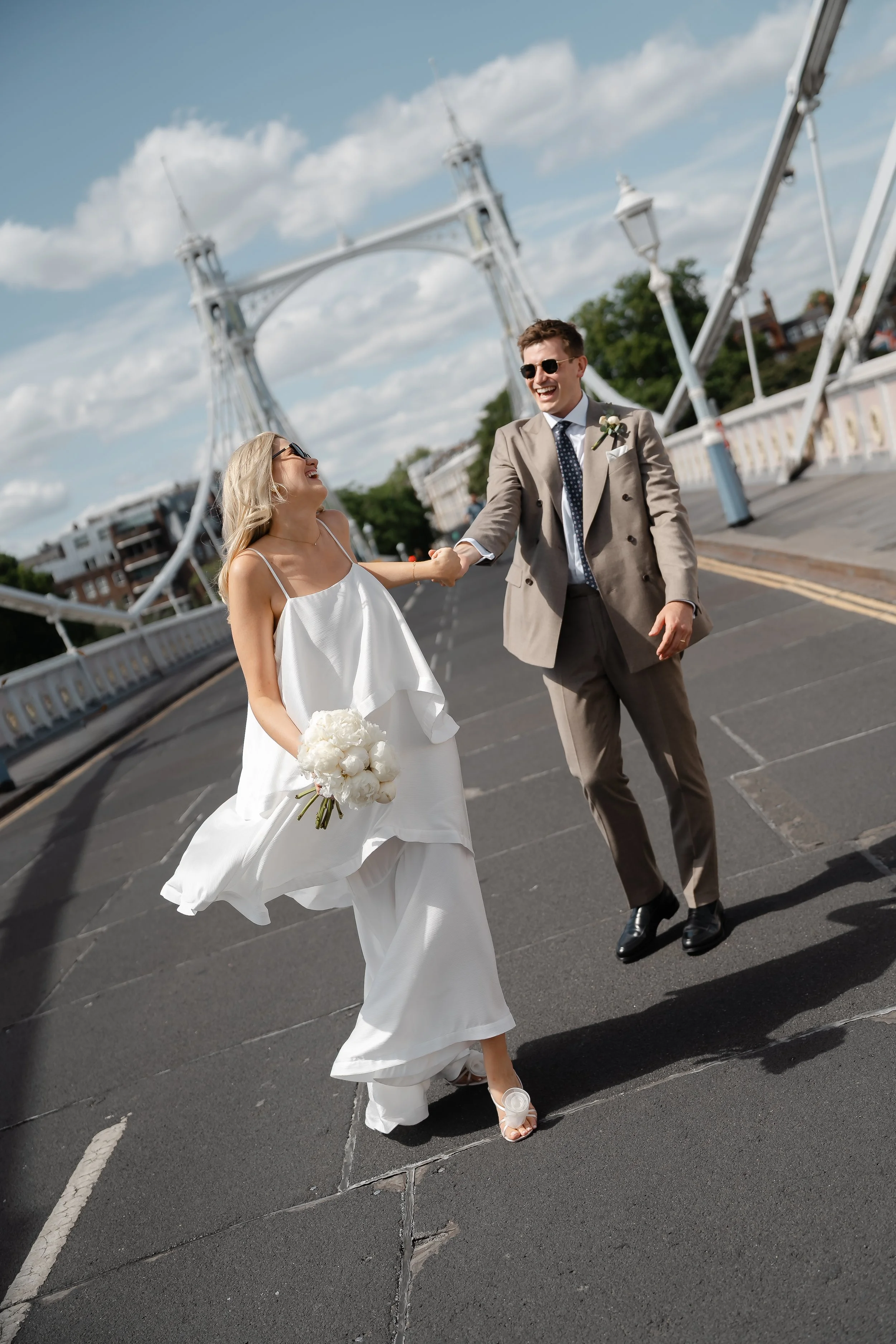 A happy bride and groom smiling and holding hands while walking on a bridge with amusement park rides in the background on a sunny day.
