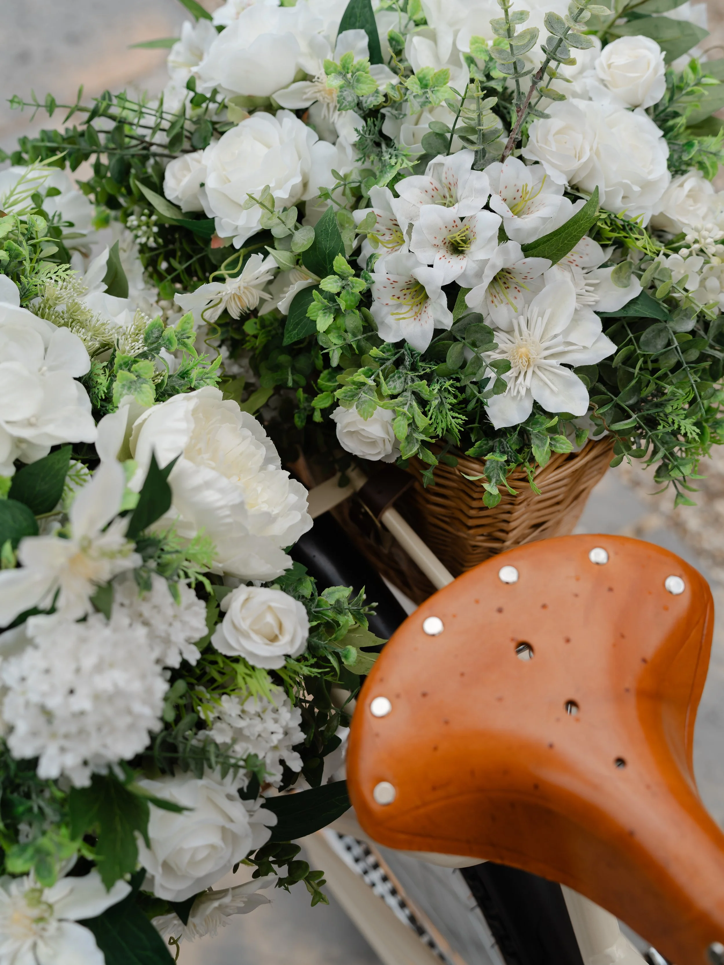 A close-up of a floral arrangement in a woven basket, including white roses, white lilies, and various green foliage, placed next to a wooden chair with circular metal accents.