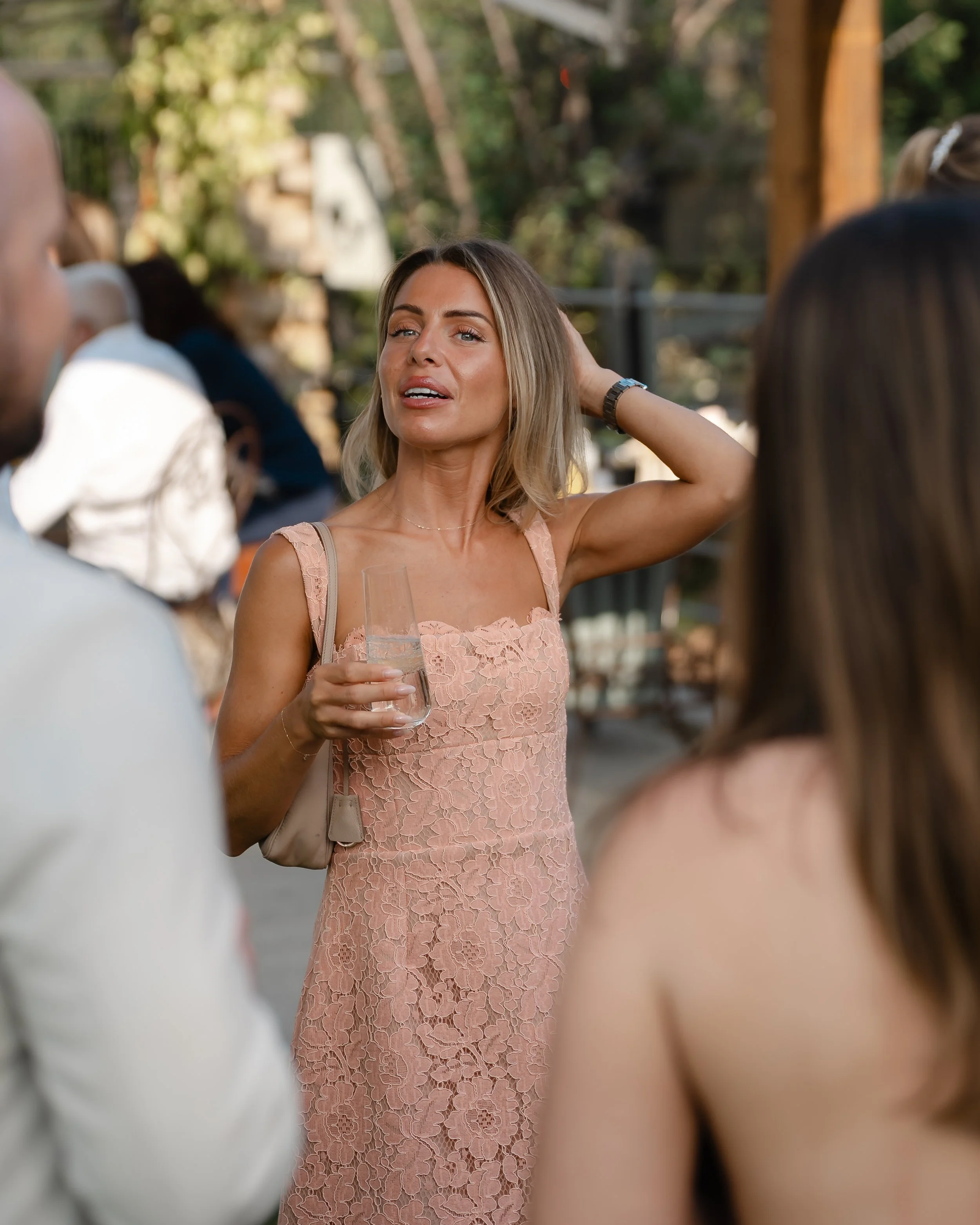 A woman in a pink lace dress at an outdoor gathering, holding a glass of champagne, with other guests and trees in the background.