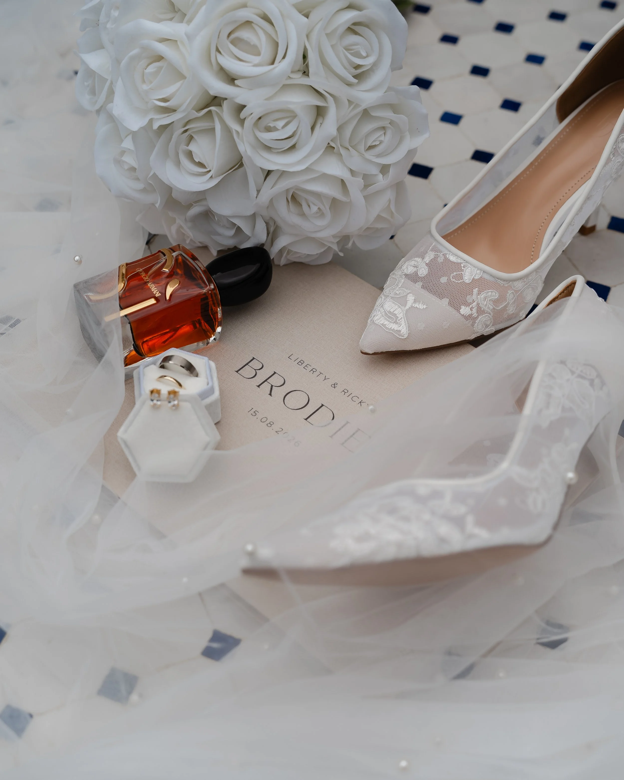 Close-up of a bouquet of white roses, a pair of white lace bridal shoes, and a bottle of perfume on a white lace tablecloth.