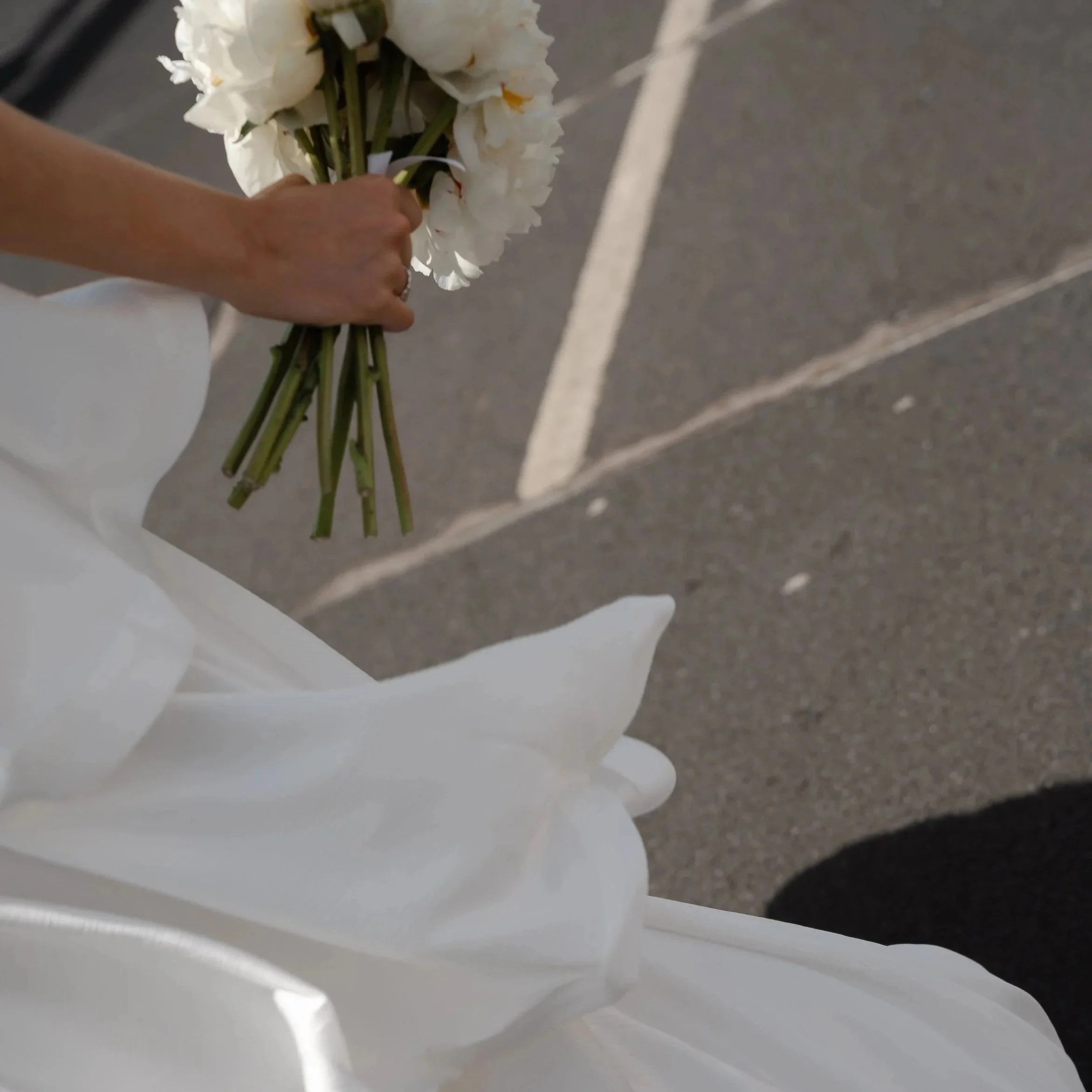 A person holding a bouquet of white flowers, possibly lilies or irises, with green stems, standing on a paved surface marked with white lines.