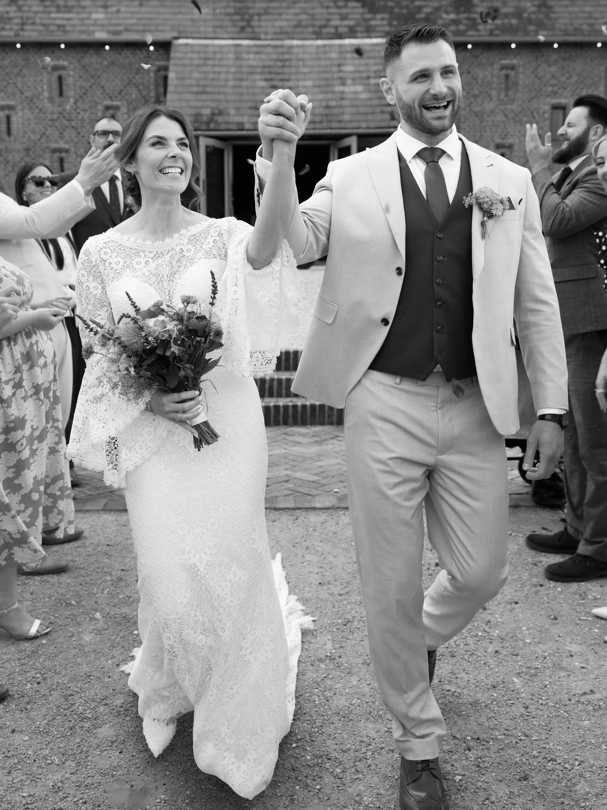 A smiling bride in a white lace wedding dress holding a bouquet and raising her hand; a groom in a light-colored suit walking beside her, holding her hand; wedding guests in formal attire celebrating around them outside in front of a brick building.