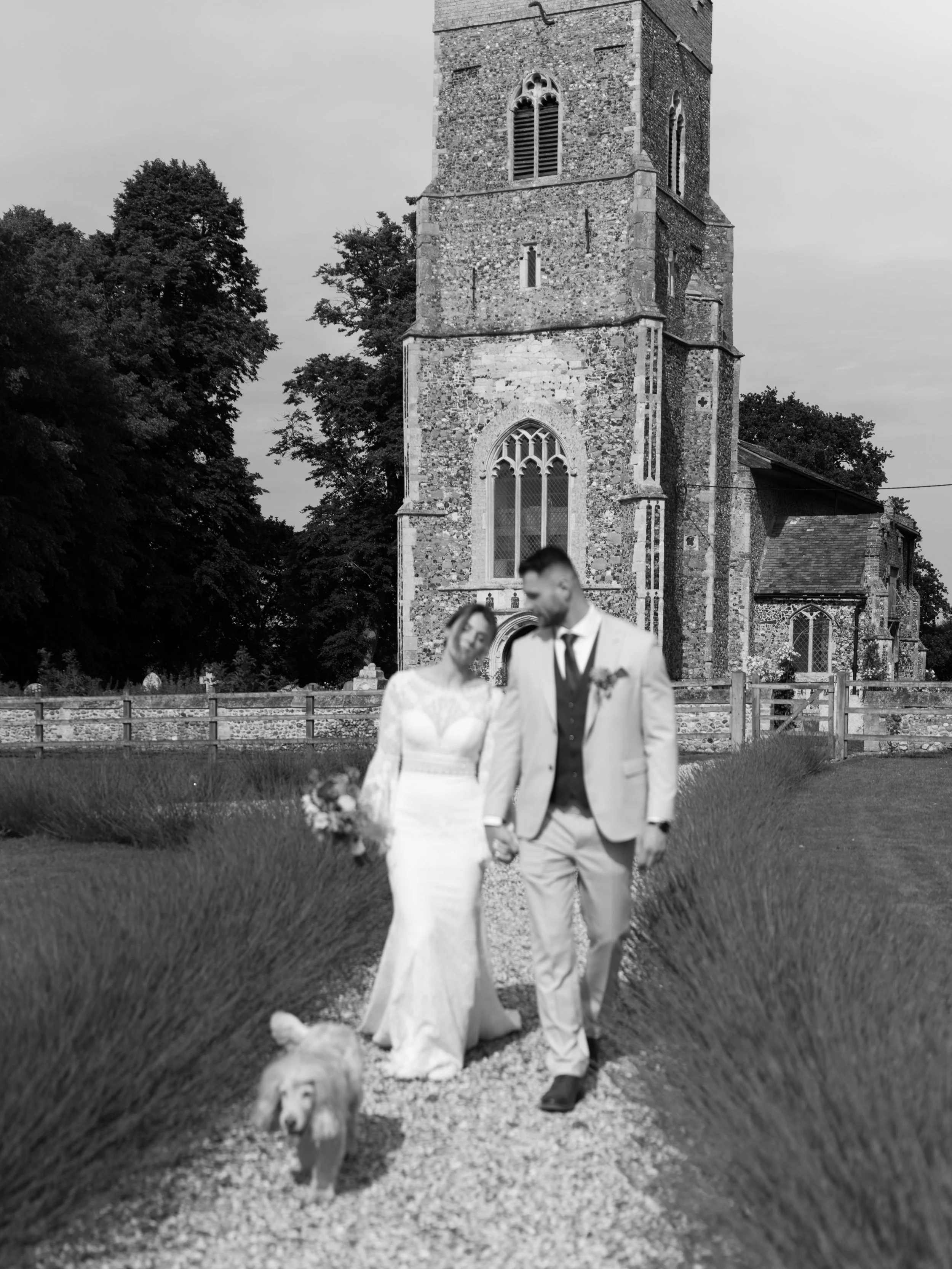 A bride and groom walking hand in hand on a gravel path near the historic stone church at Copdock Hall, with tall, pointed arch windows and a clock tower, surrounded by trees.