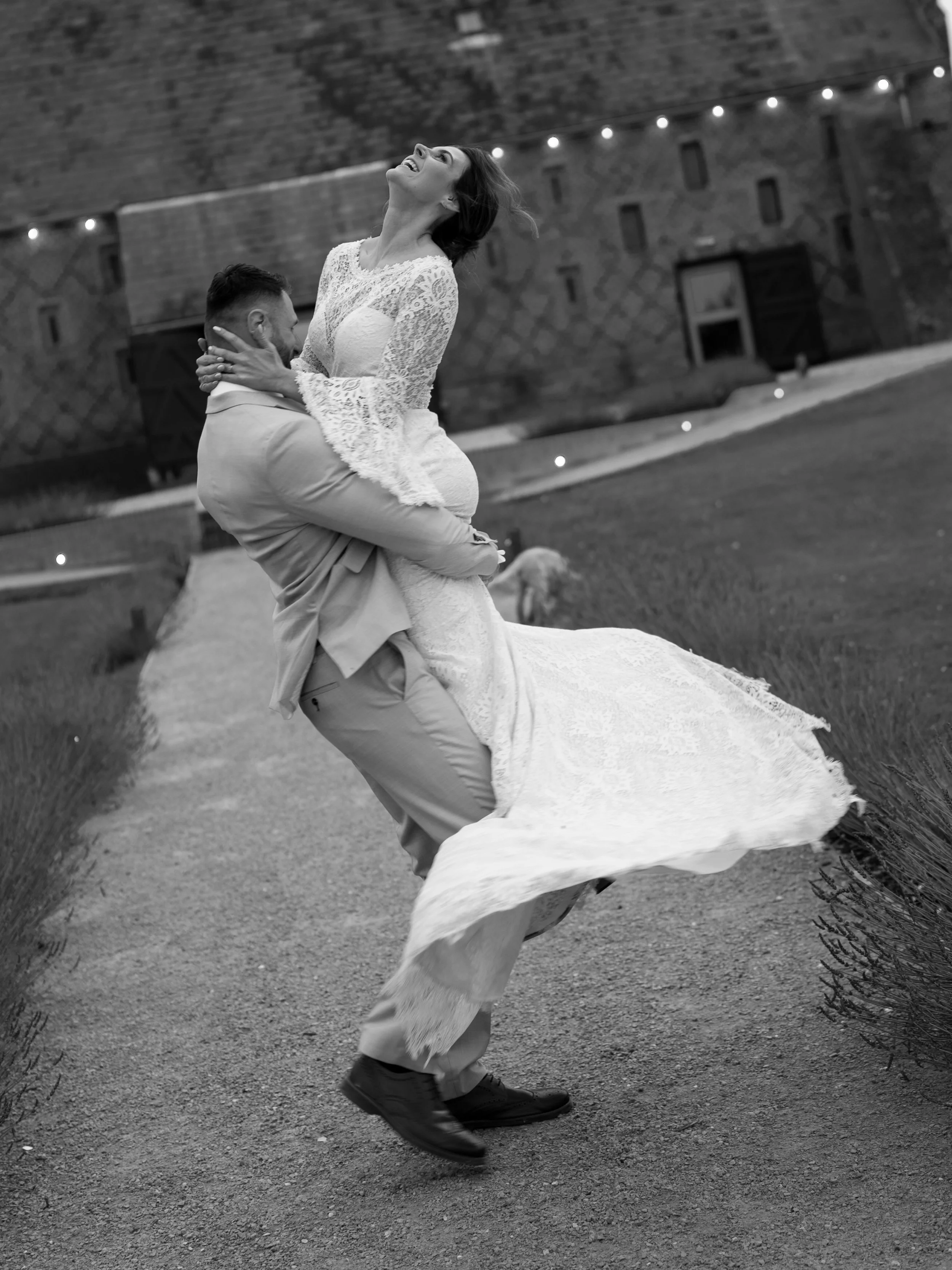 A black and white photo of a man lifting a woman in a wedding dress outside at night. The woman is laughing and the man is smiling, with a brick building and string lights in the background.