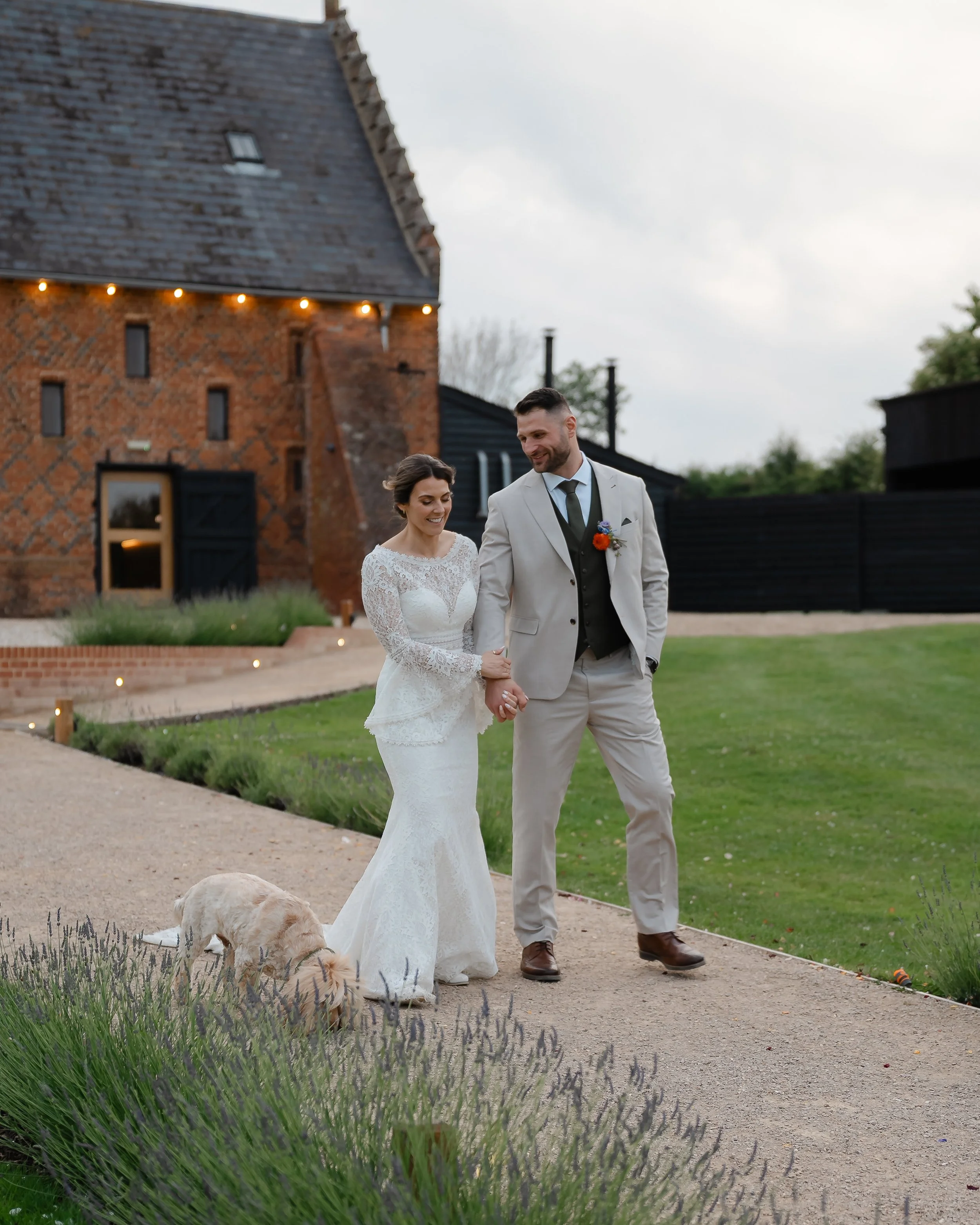 A bride and groom walking hand in hand outdoors, smiling, with a dog in front of them, in front of a the Edwardian barn at copdock hall, suffolk, with string lights and a cloudy sky.