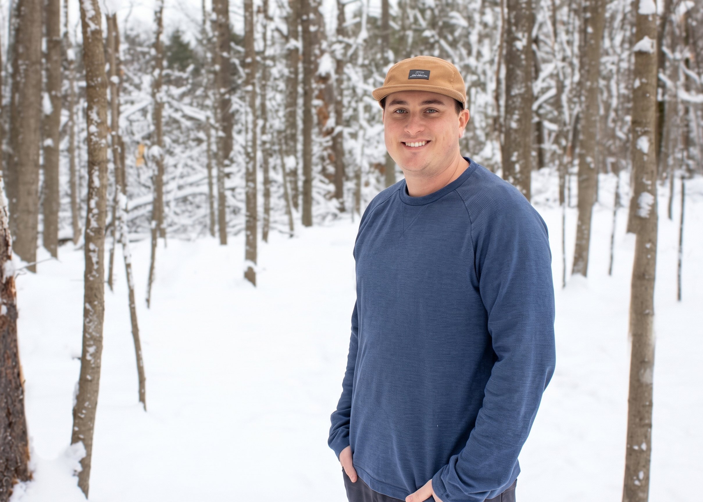A young man in a blue long-sleeve shirt and tan cap standing in a snowy forest with tall trees.