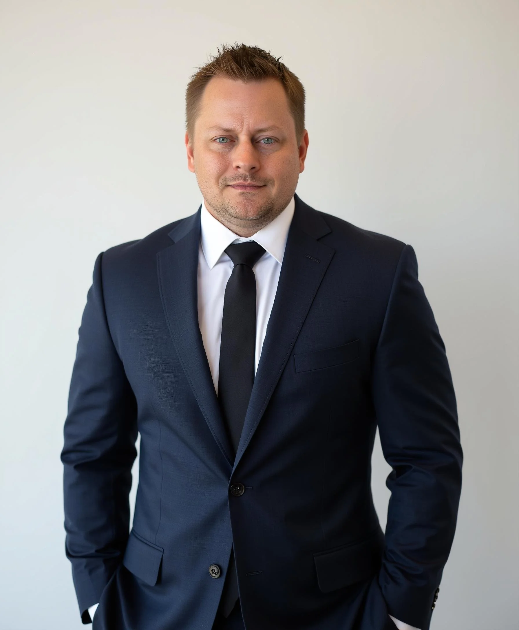 Man in a dark suit with a white shirt and black tie standing against a plain background.