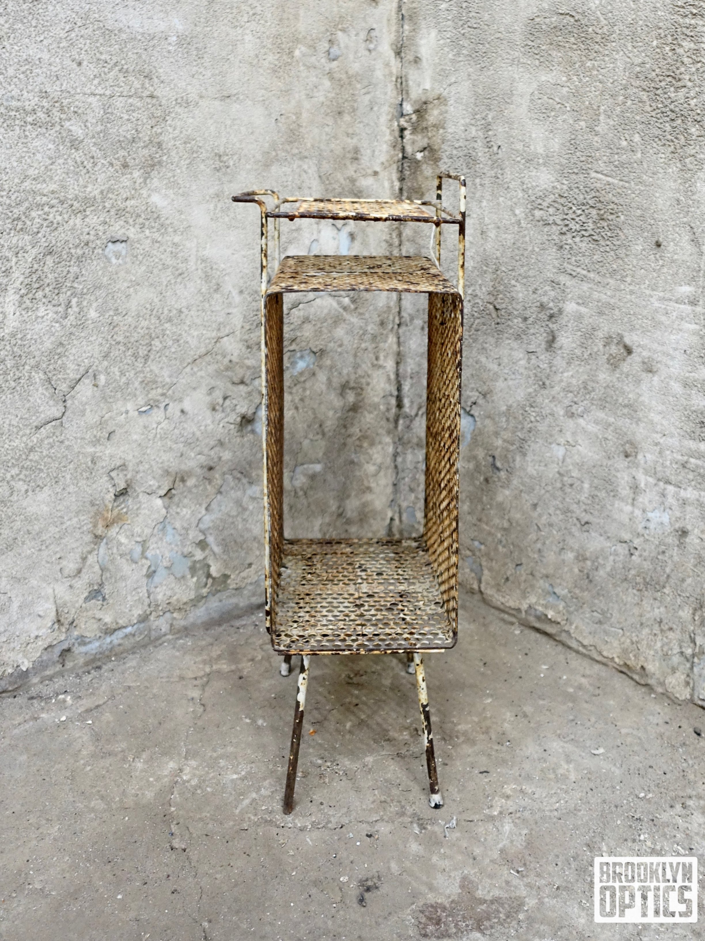 A rusty, small, three-tier metal shelving unit with a lattice design, standing on three thin, uneven legs against a rough, gray, concrete wall.