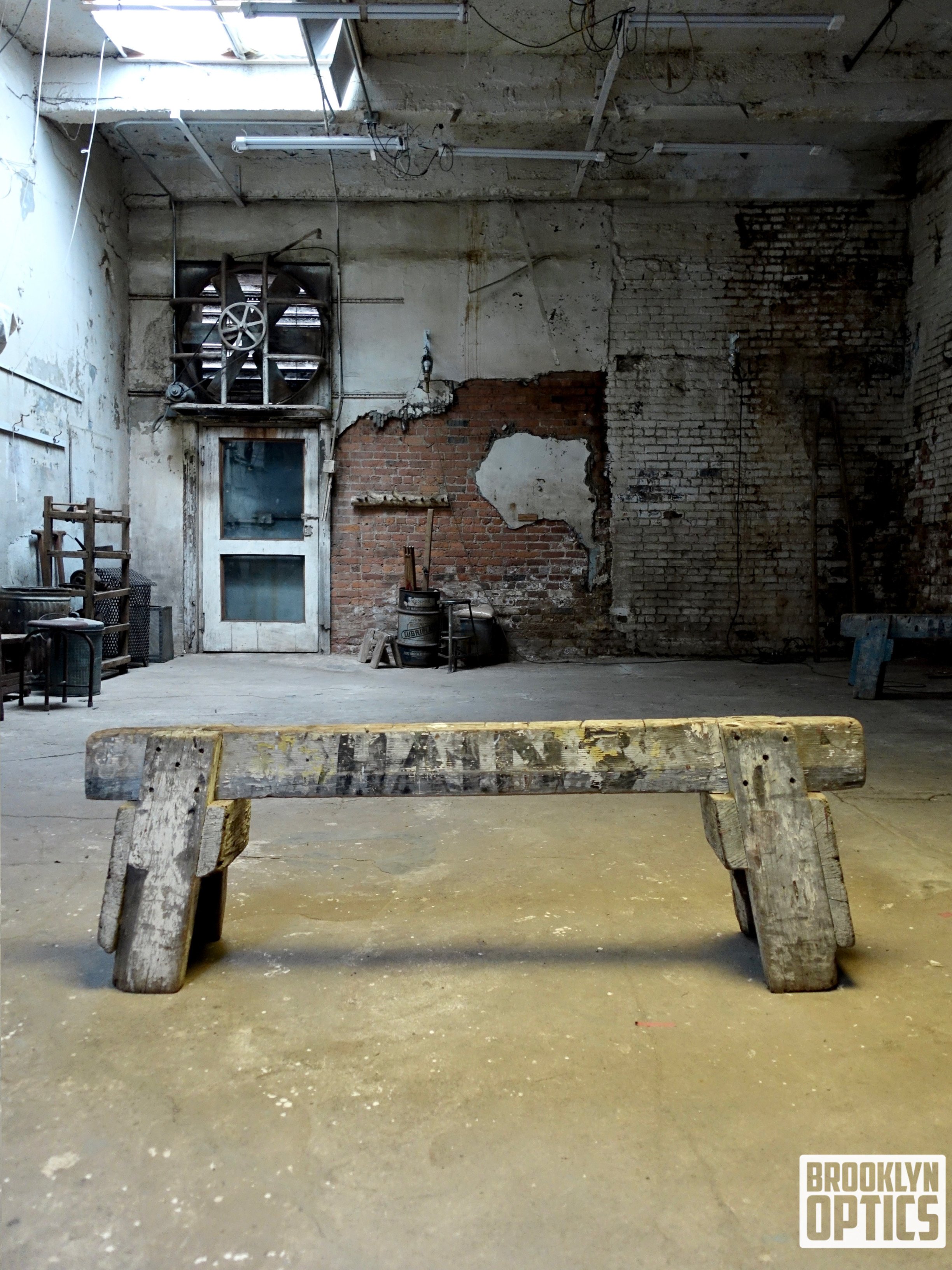 Old, weathered wooden bench in an abandoned, industrial space with exposed brick walls and peeling paint, Brooklyn Optics logo in the bottom right corner.