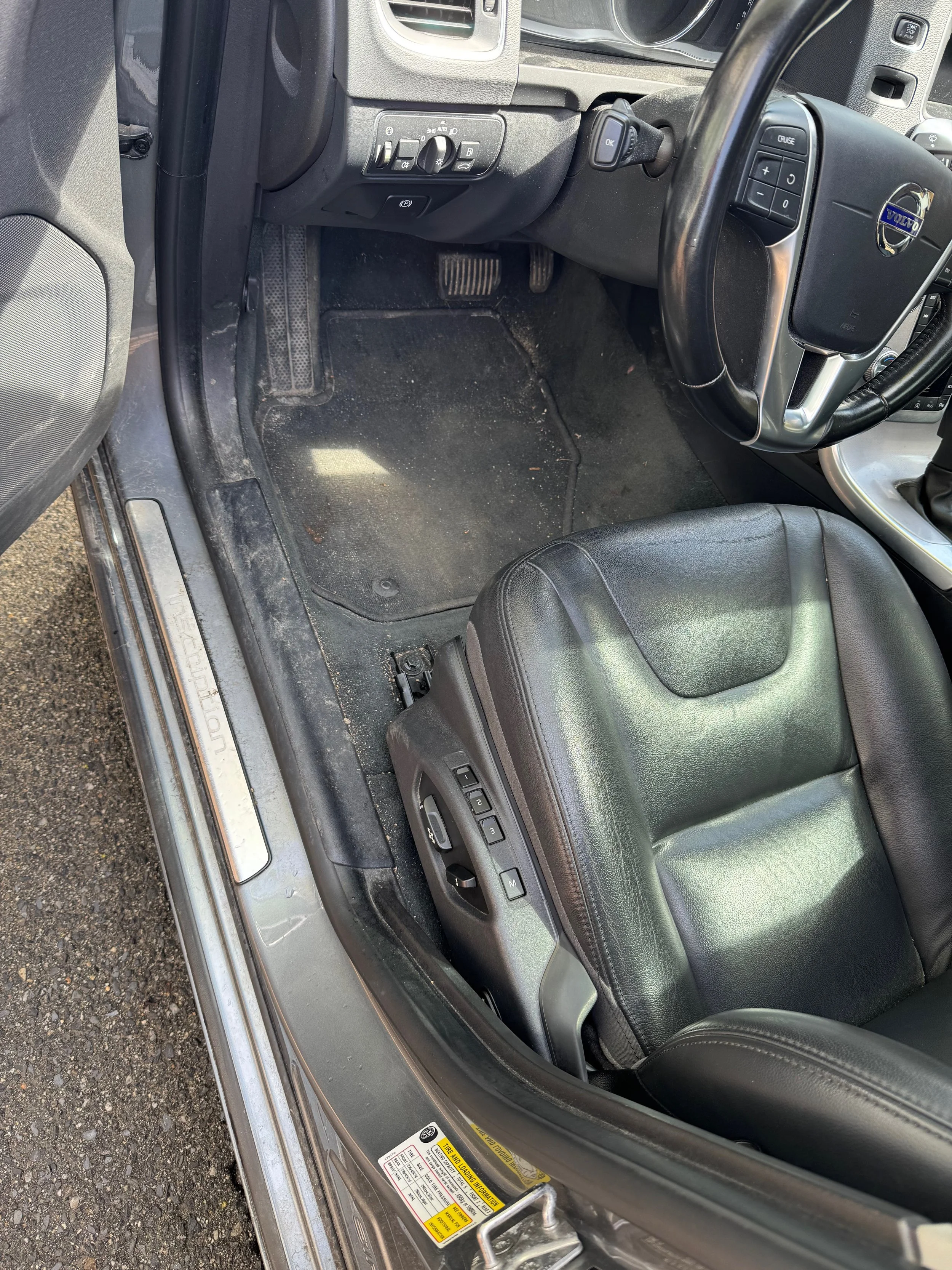 Inside view of the driver's side of a black Volvo car showing the black leather seat, steering wheel, dashboard, and footwell area with a dirty floor mat.
