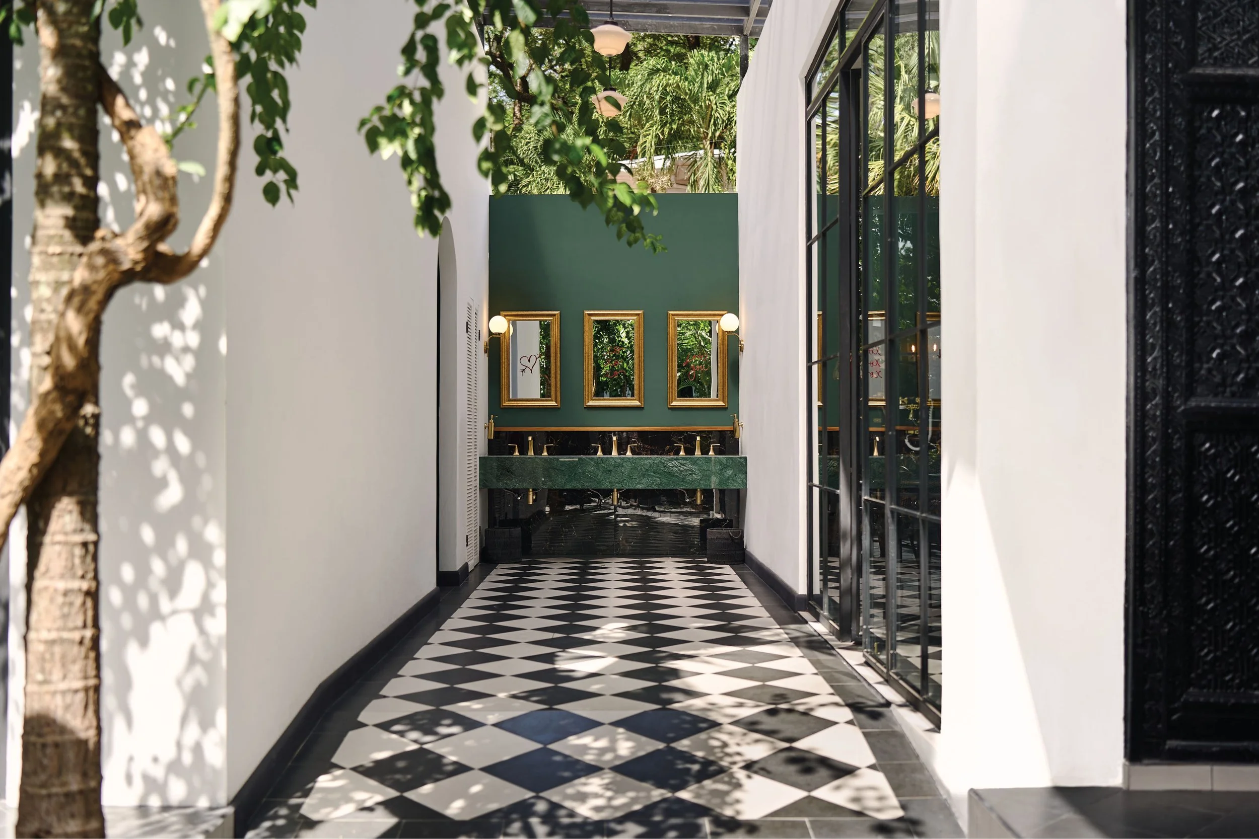 A narrow hallway with black and white checkered flooring, white walls, large glass doors on the right, and a green wall with three gold-framed mirrors and two wall lamps on the far end. Sunlight and shadows from trees are visible.