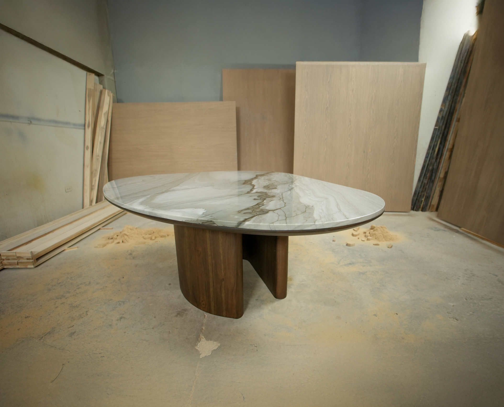 A modern table with a marble top and wooden base, in a workshop with large wooden panels and construction materials in the background.