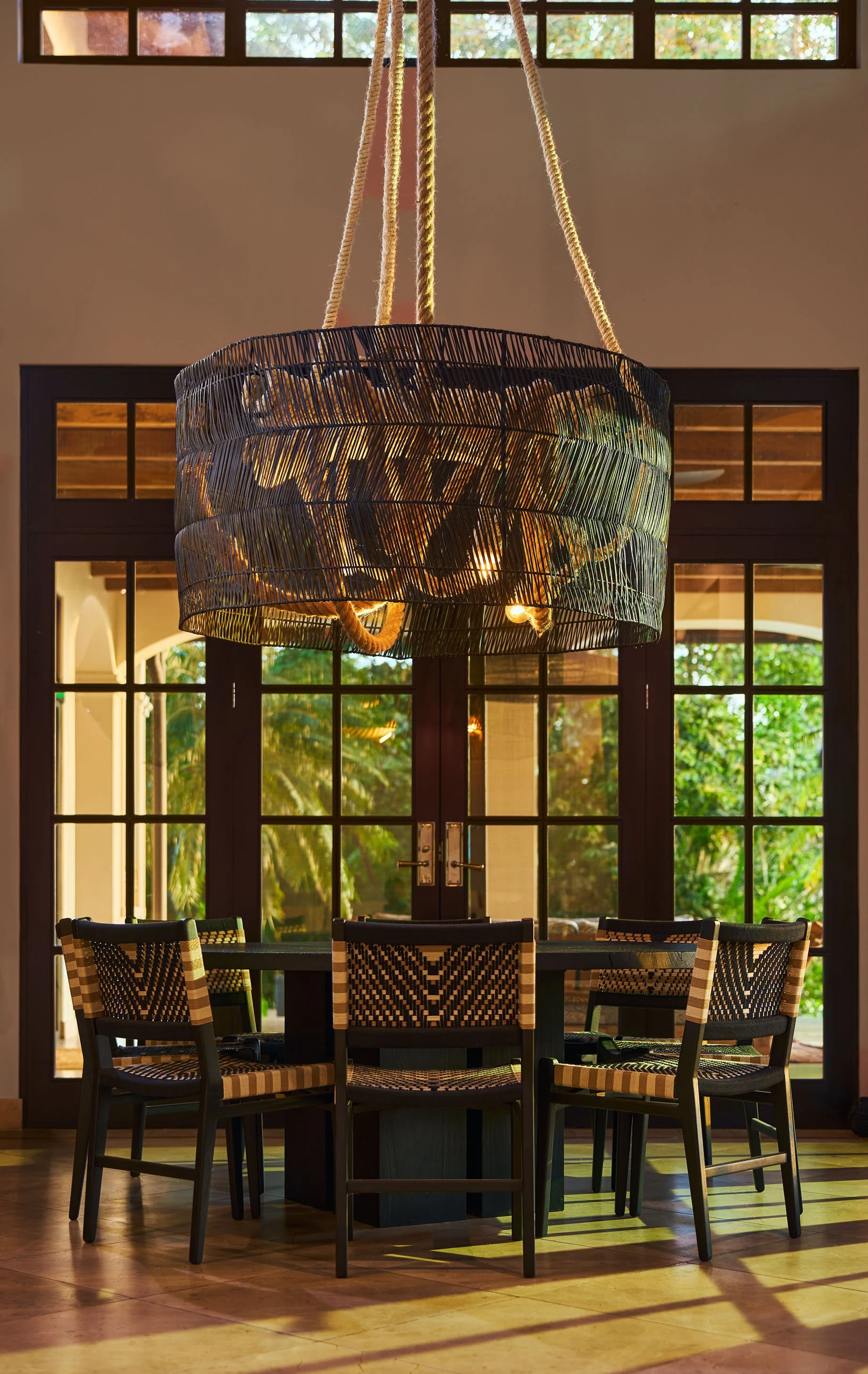 Woven pendant lamp in front of arched steel-framed windows facing the jungle
