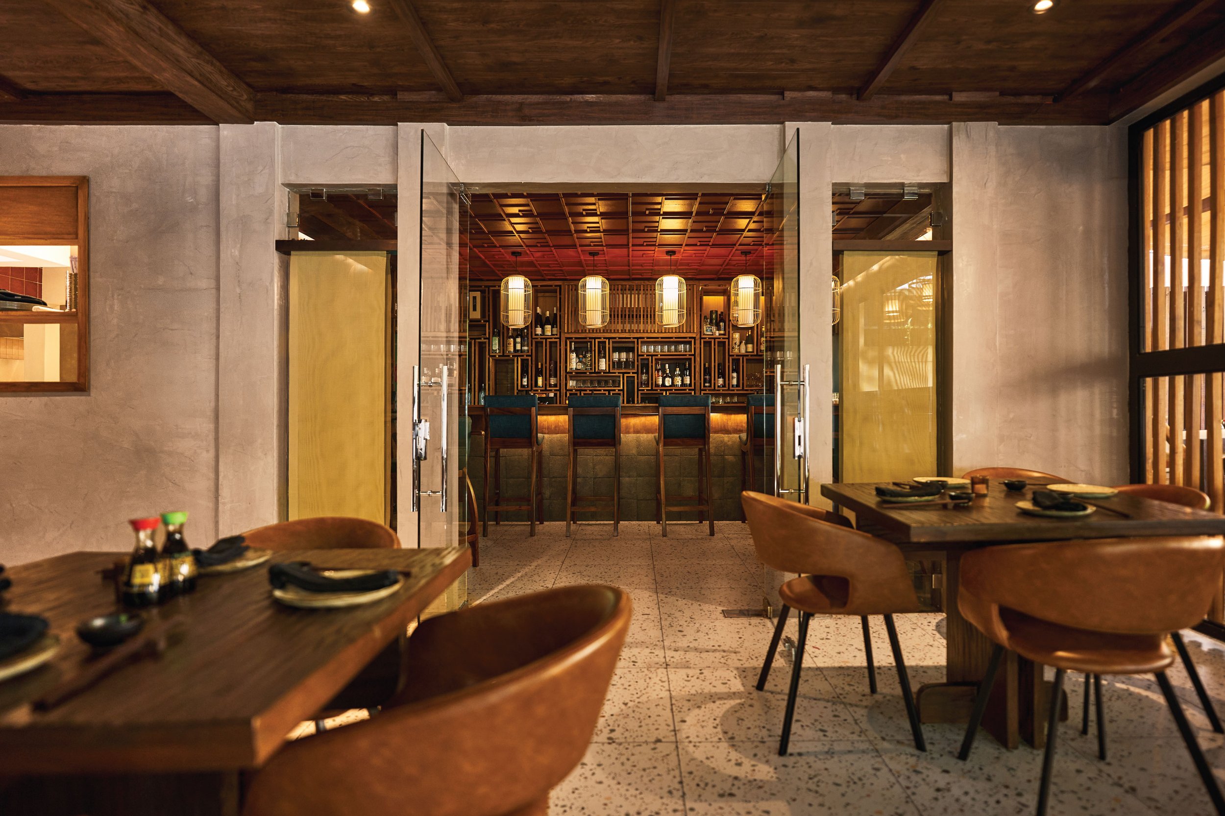 Interior of a restaurant dining area with wooden tables and chairs, a bar with bottles and glasses, decorative lantern lights, and Japanese-style sliding doors.