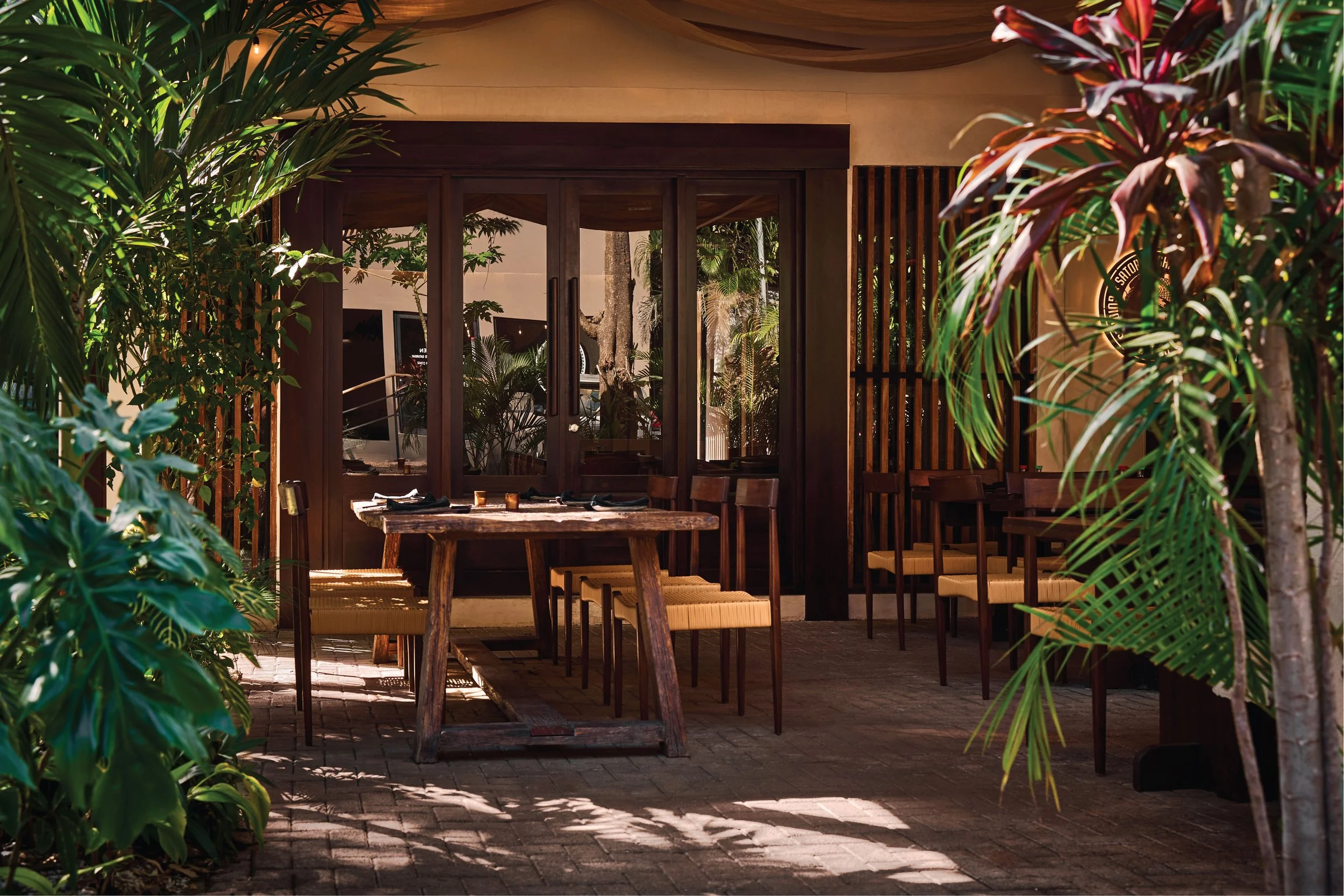 Interior of a lush, green restaurant or cafe with wooden furniture, sunny natural light, and hanging plants.