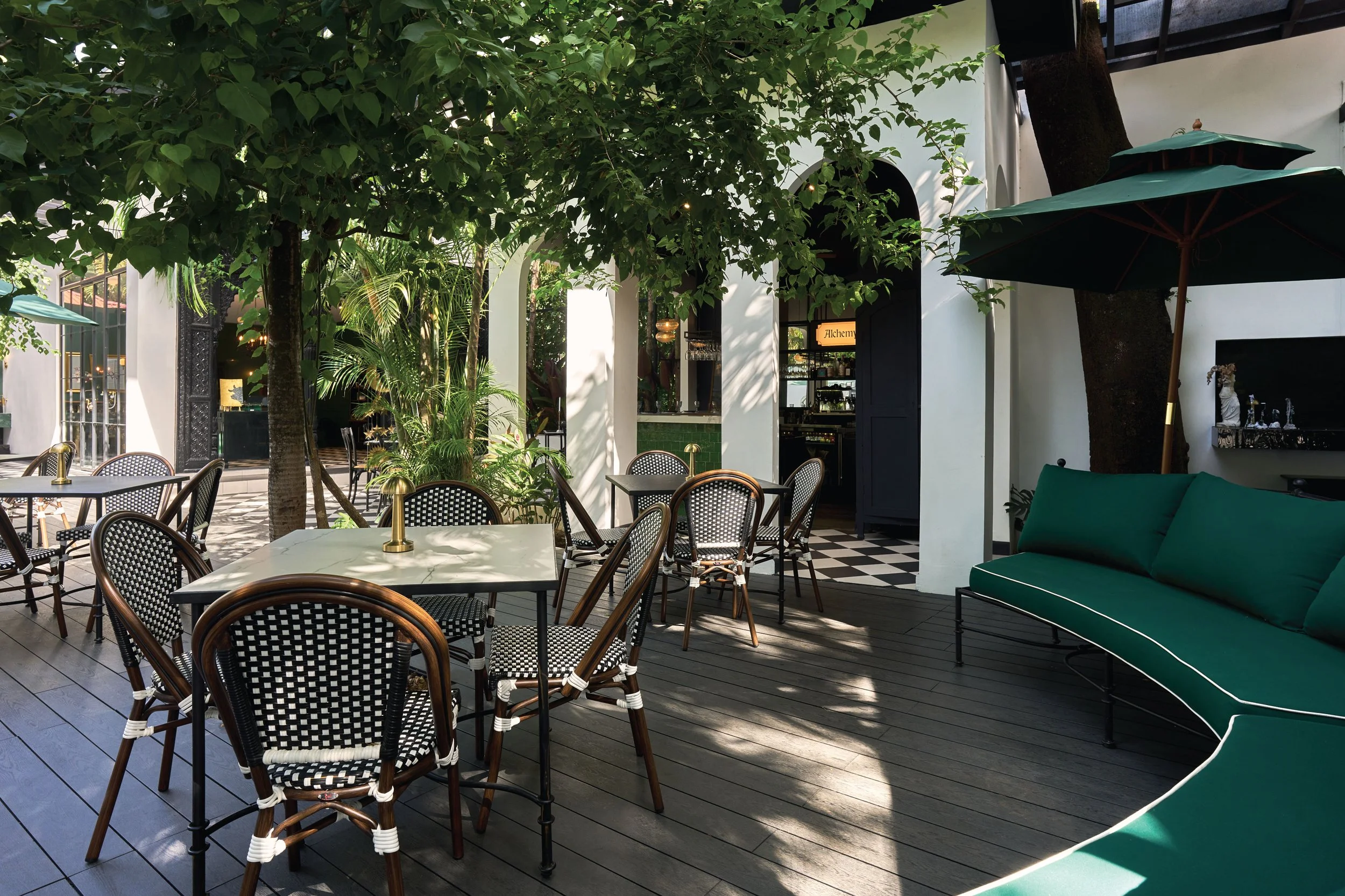An outdoor patio with black and white checkered flooring, round tables with black and white chairs, green umbrellas, and lush green trees. There is a building with large windows and open doors, and some interior decor is visible.