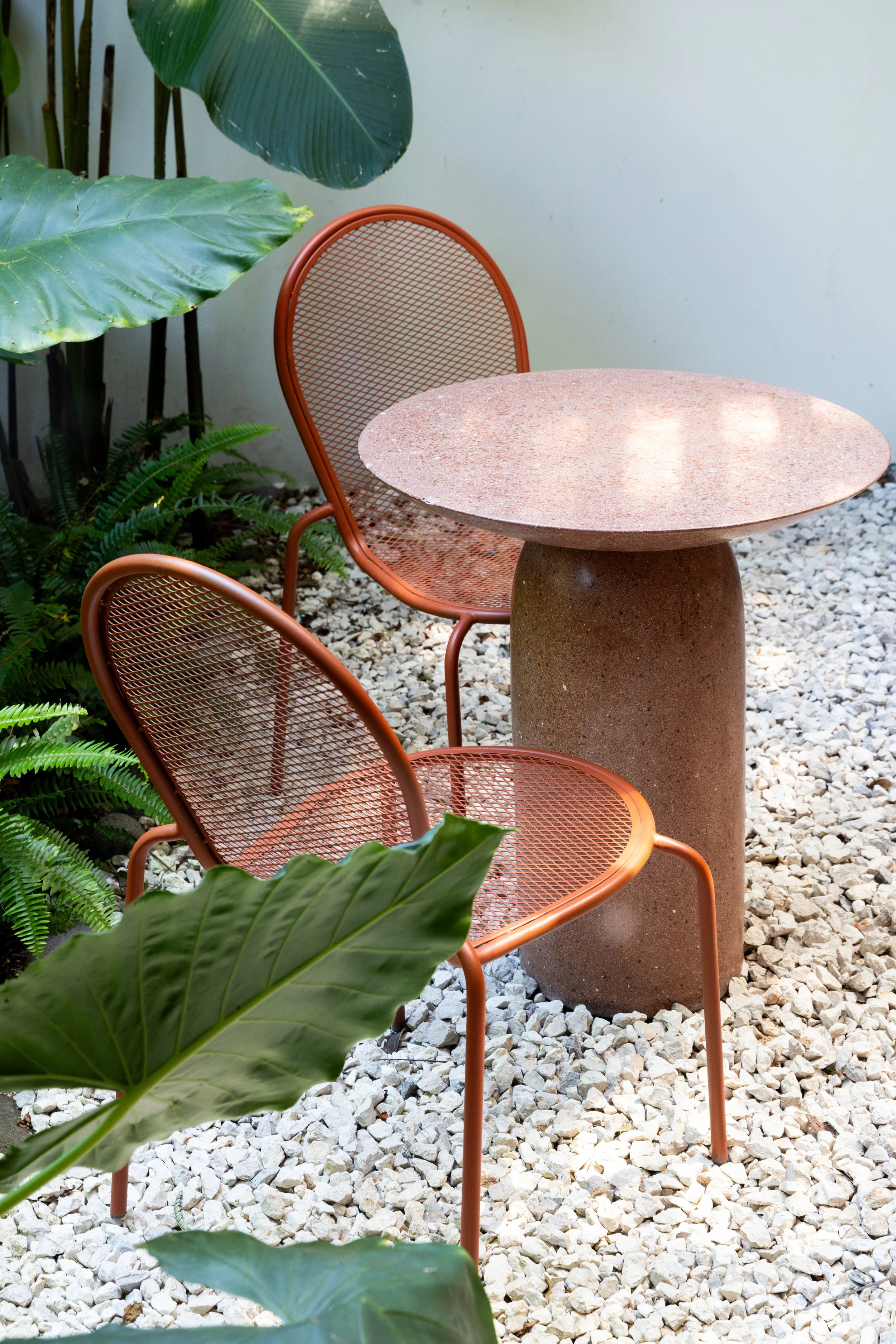 Two orange metal chairs and a round stone table in an outdoor setting, surrounded by green tropical plants and white gravel ground.