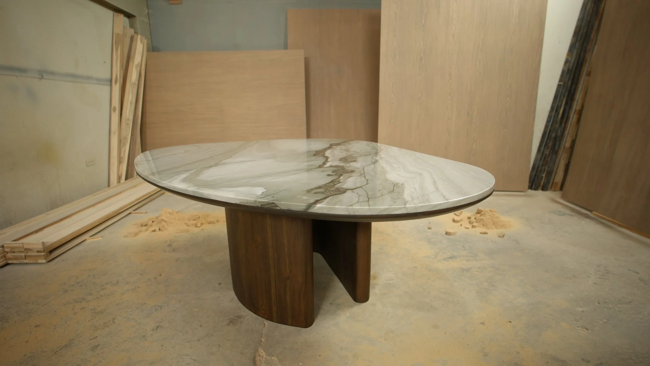 A modern table with a marble top and wooden base in a workshop setting, surrounded by wood panels and construction materials.