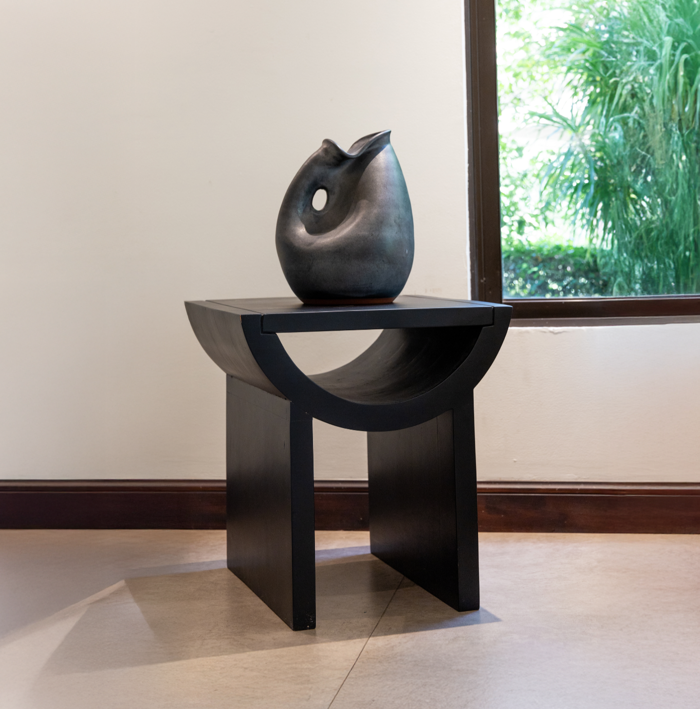 A black ceramic vase with an abstract, curved opening, placed on a modern black wooden pedestal near a window with green foliage outside.