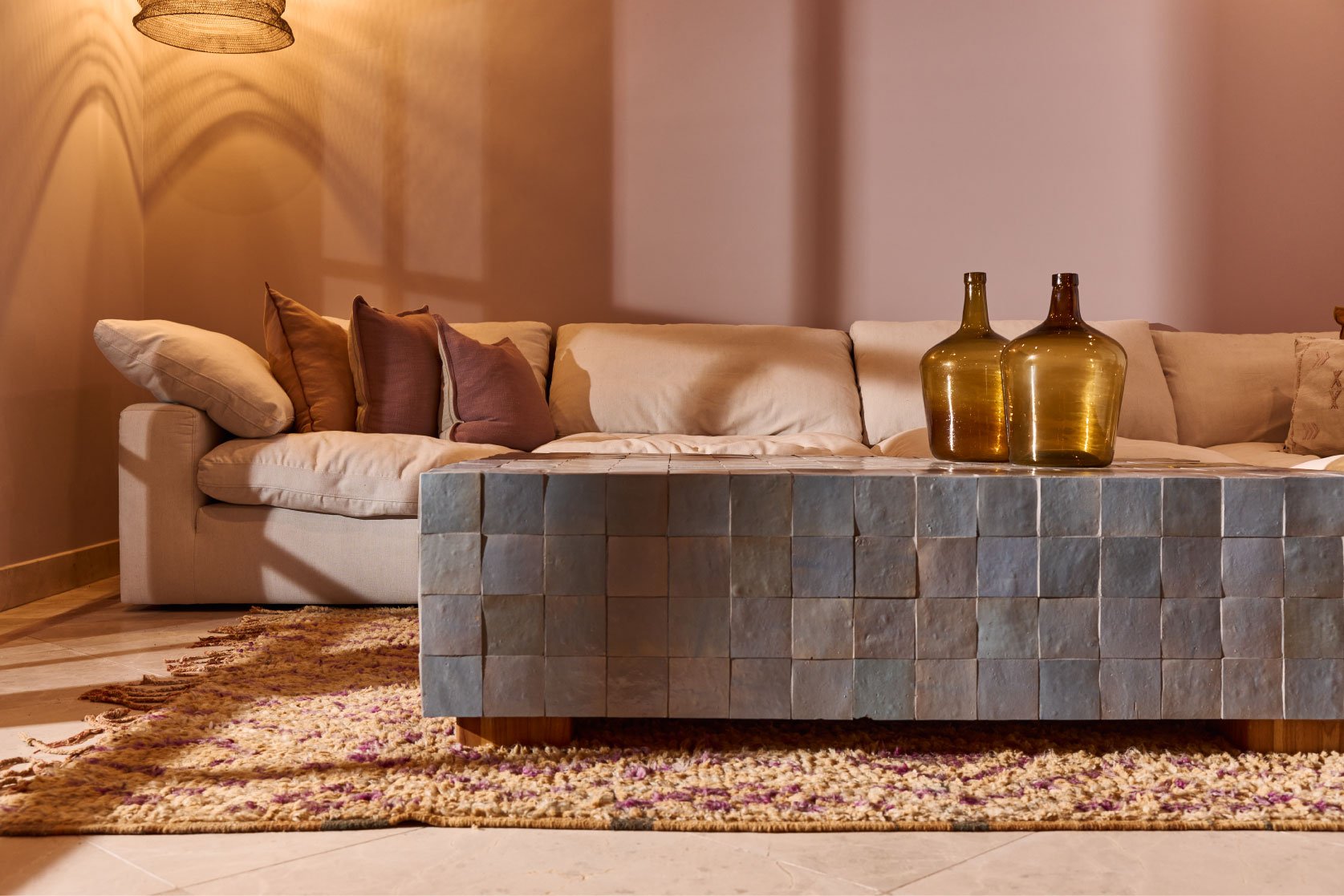 A beige couch with multiple brown and beige pillows, placed in front of a plain wall. Two amber-colored glass vases are on a tiled coffee table in front of the couch, with a multicolored woven rug underneath.