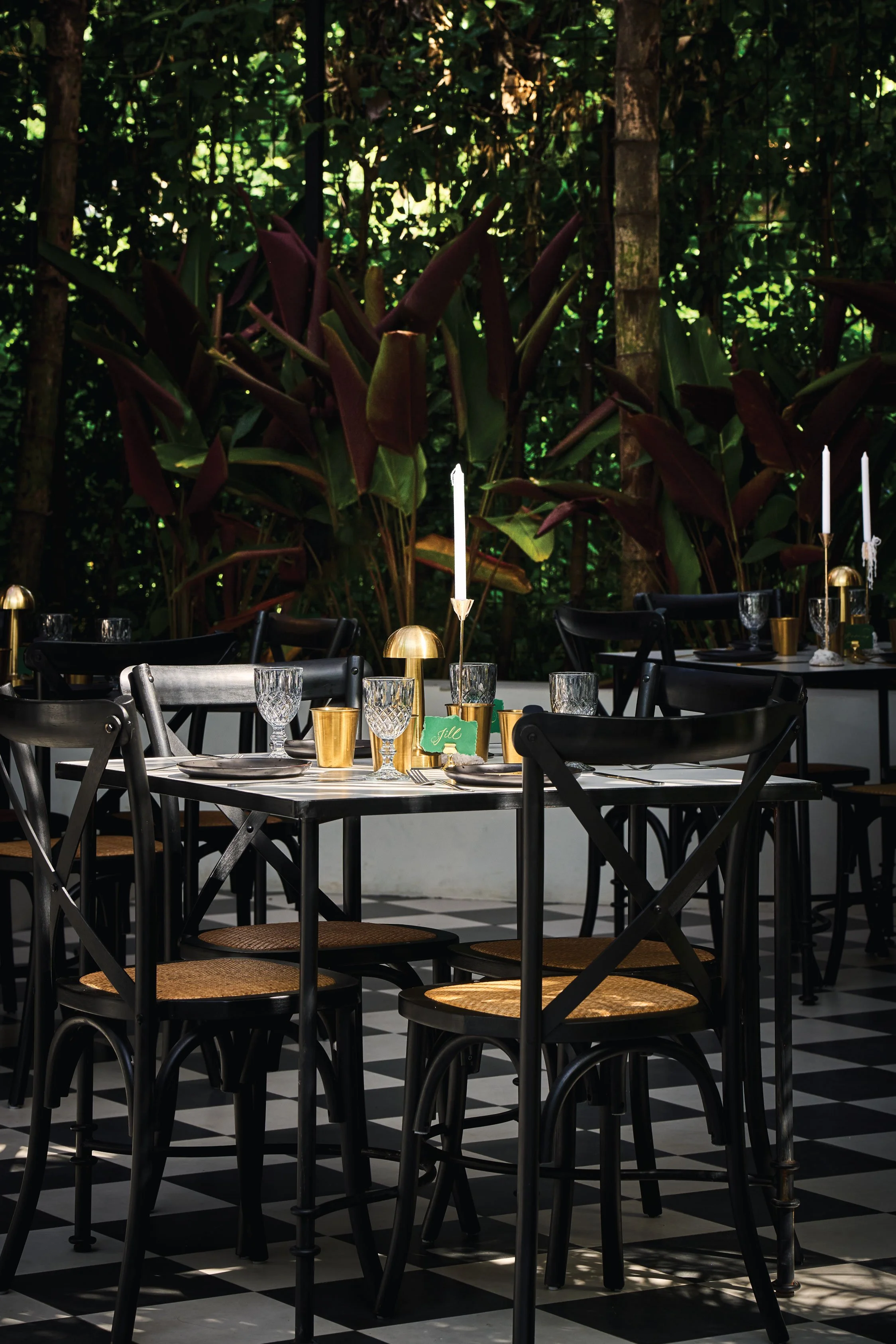Interior of a restaurant with black chairs, a checkered black and white floor, tables set with glassware, gold candles, a green place card, and a lush green outdoor garden backdrop visible through large windows.
