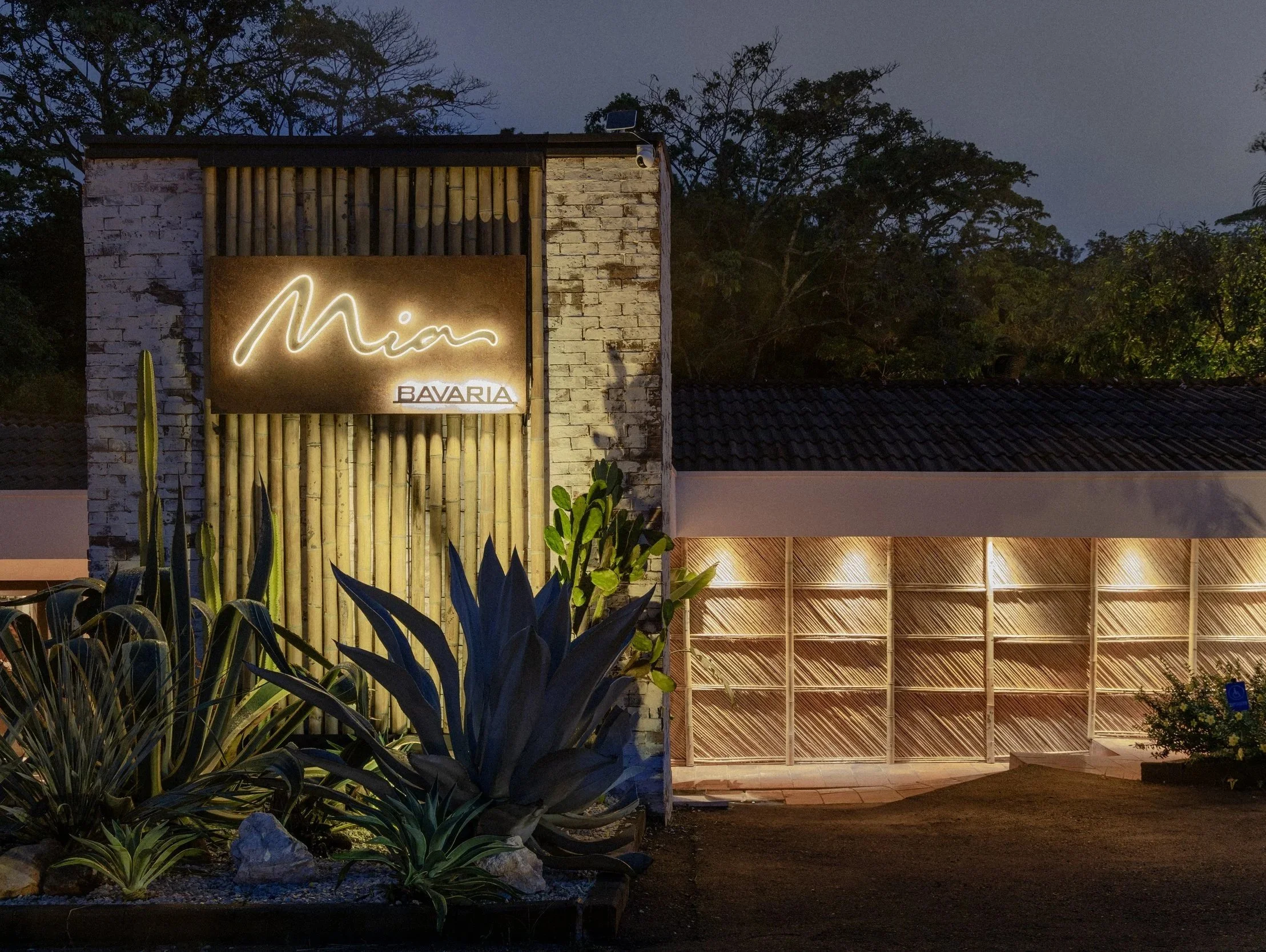 Nighttime exterior view of a building with a lit neon sign reading 'Mina' and smaller text 'BAVARIA'. The building has a brick wall, bamboo decor, and a landscaped area with succulents and tall cacti in front. There is warm lighting illuminating part of the building's garage or entrance area.