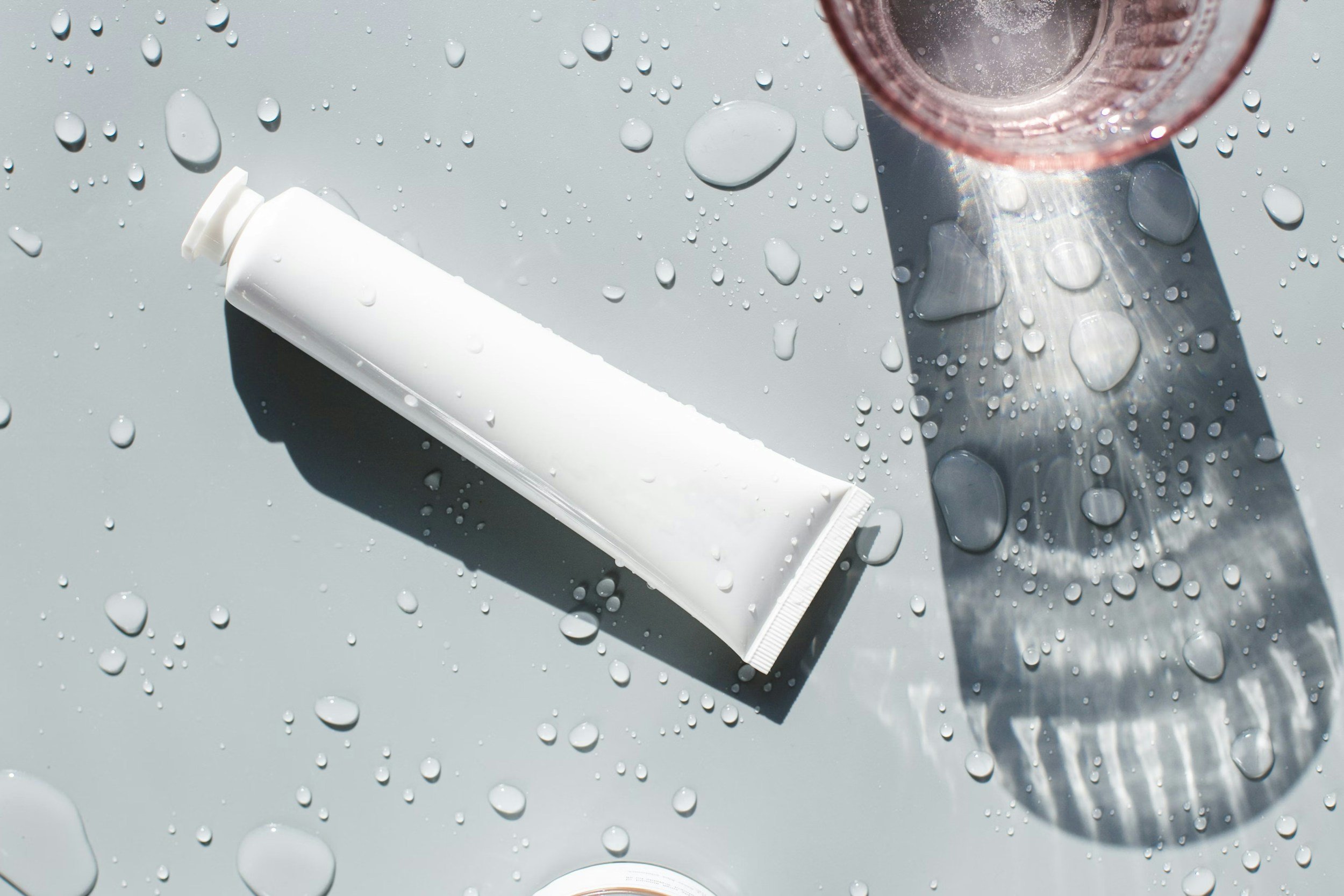 White toothpaste tube lying on a wet surface with water droplets and sunlight reflections.