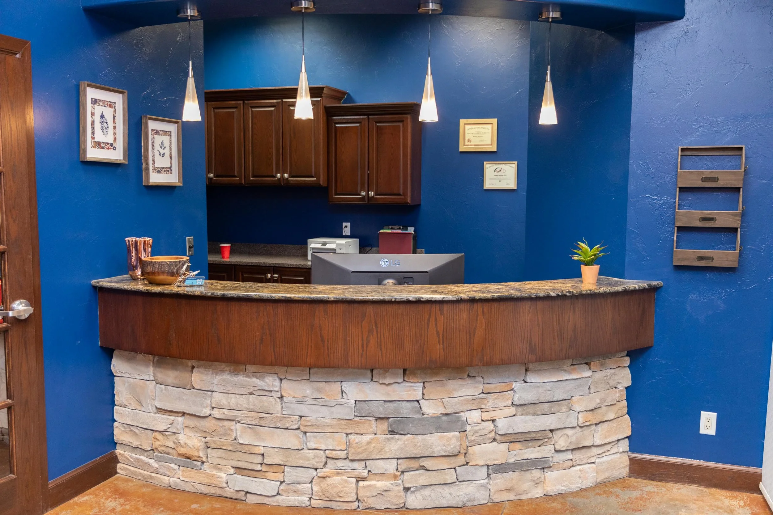 Interior of a reception area with a curved stone and wood counter, blue walls, three hanging pendant lights, framed certificates, and decorative items including a small potted plant and bowls.