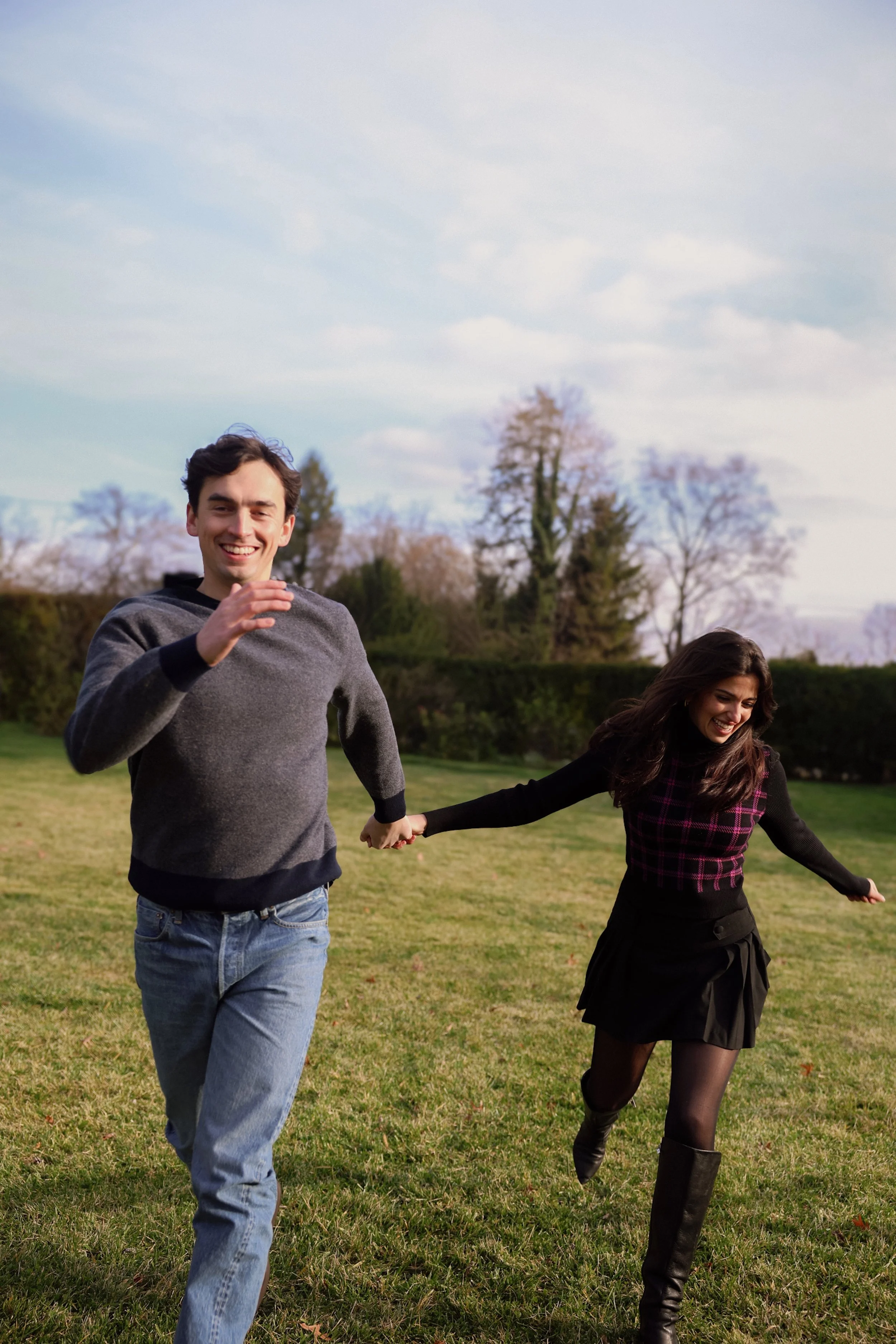 A young man and woman are running and playing together outdoors on a grassy field, holding hands, with trees and cloudy sky in the background.