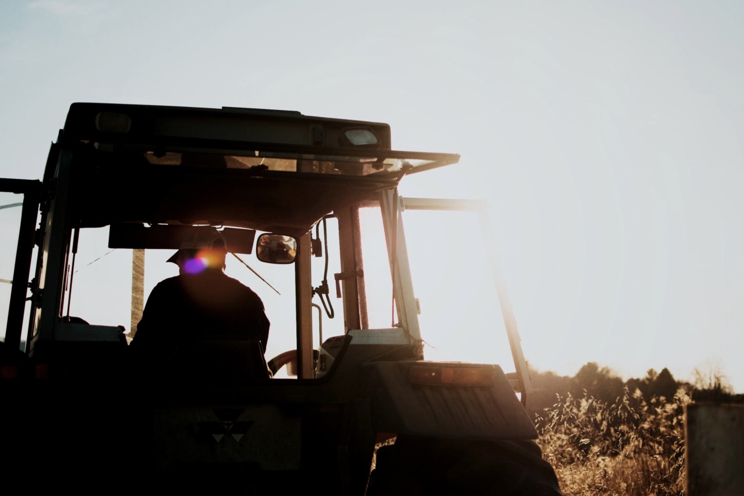 Farmer operating tractor on a farm at sunrise or sunset, with sunlight glowing behind the vehicle.