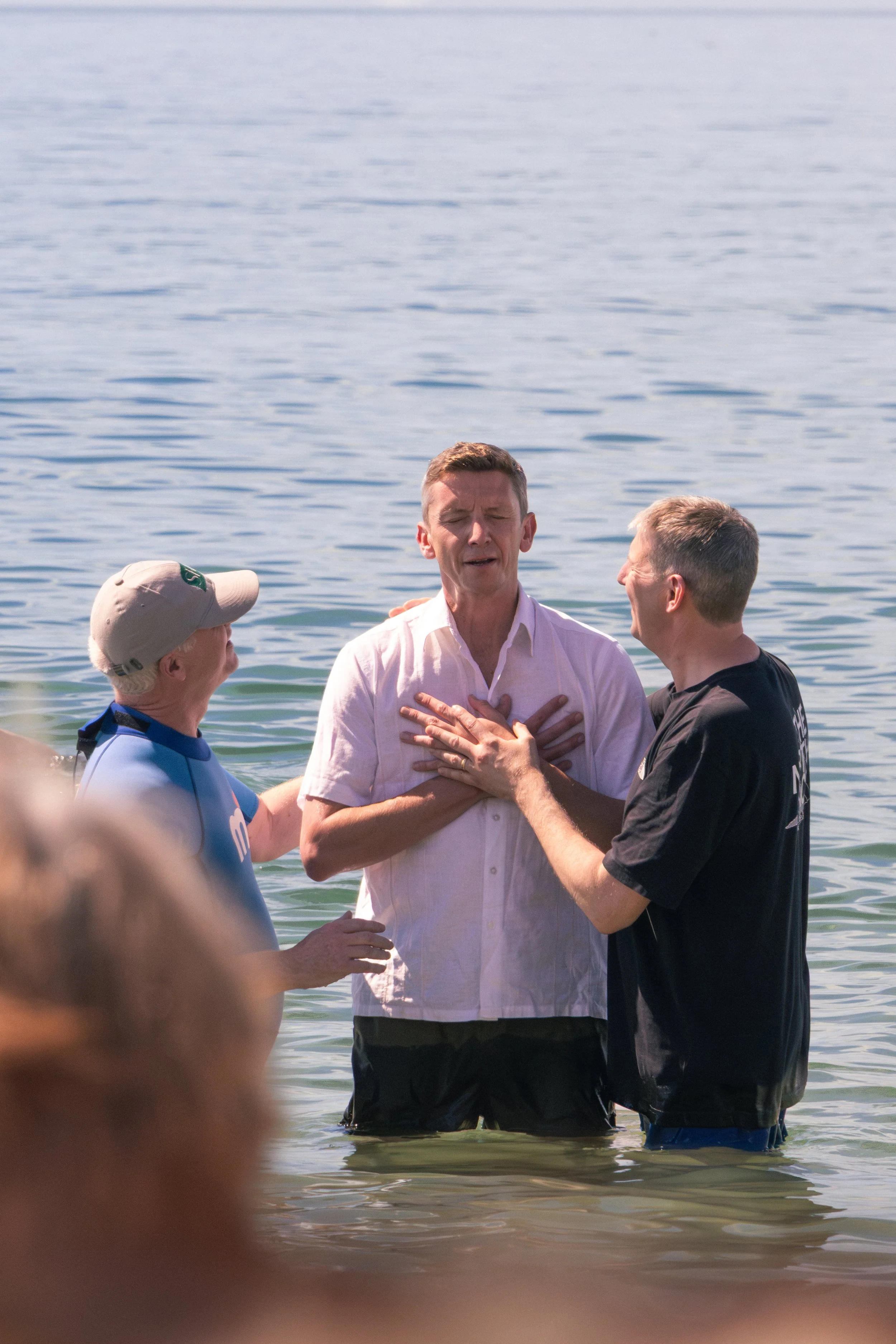3 men standing in water, with one man at the center clutching his chest and eyes closed, while others reach out about to baptise him. The background shows a body of water.