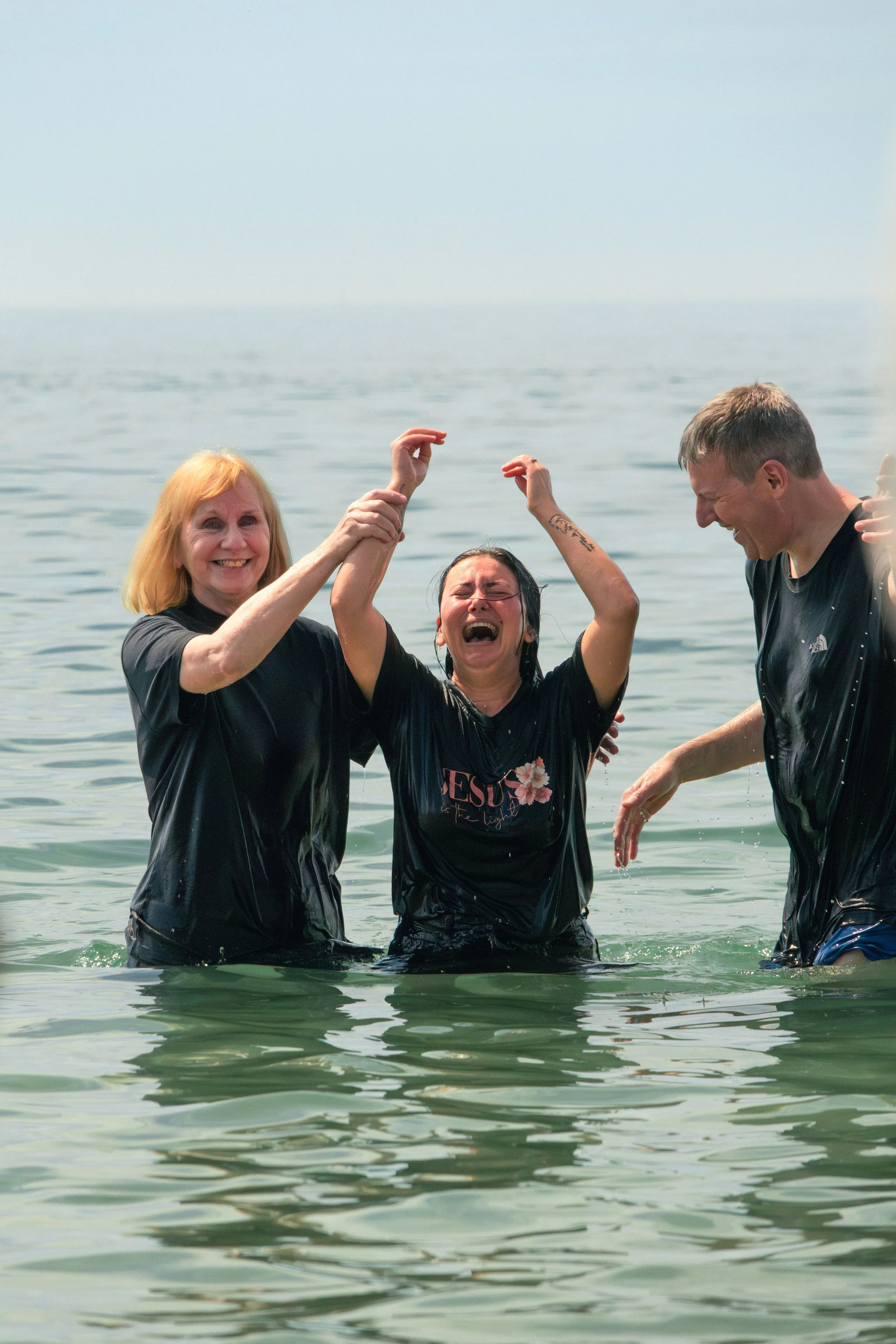 Three people standing in water, holding hands, smiling and laughing in an outdoor setting, with a calm body of water behind them as one person has been baptised in the middle. Her hands are raised in joy and exclamation.