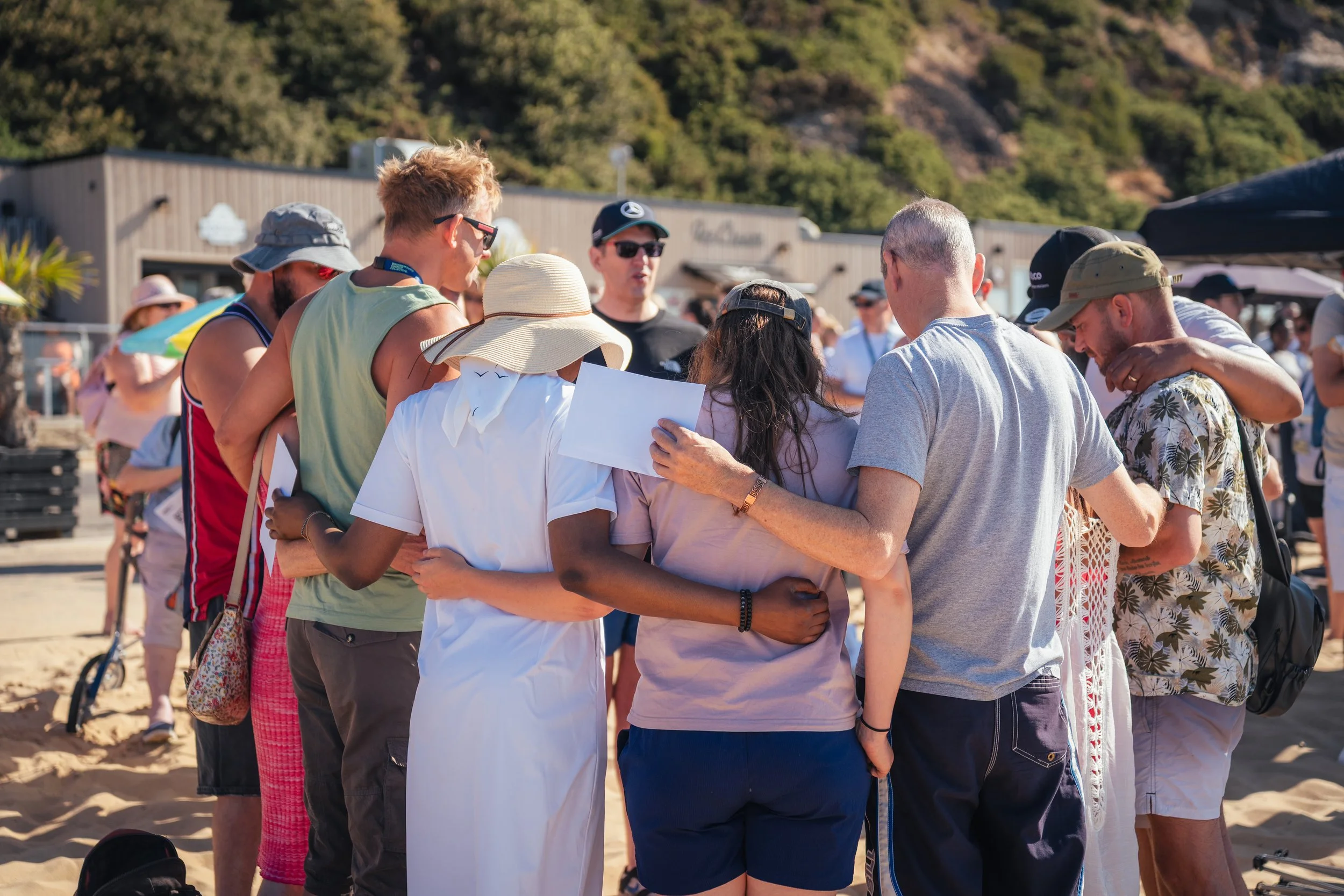 A group of people embracing each other on a beach during daytime, with some wearing hats and casual summer clothing.