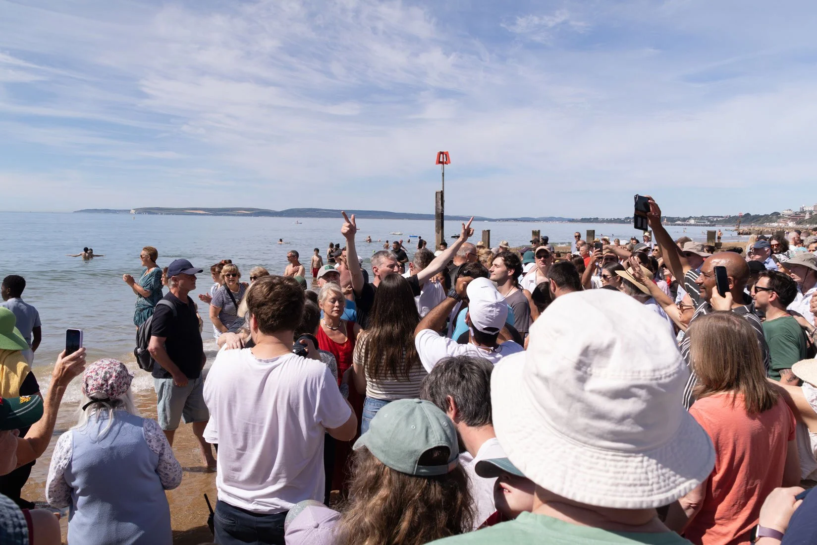 Crowd of people on a beach with some in the water, some taking pictures, and a person making a peace sign in the centre, at a Beach Baptisms.