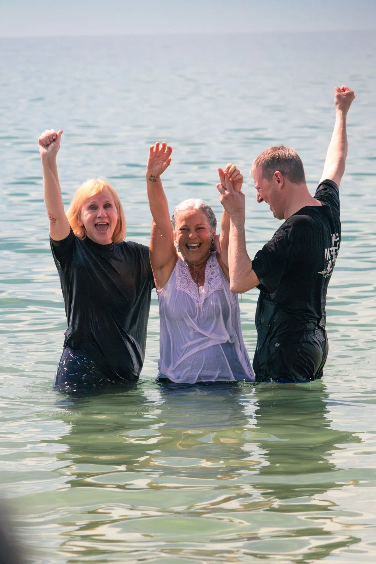Three smiling people, two women and one man, celebrating in the sea with their arms raised after the woman was baptised, during daytime at Boscombe Beach.
