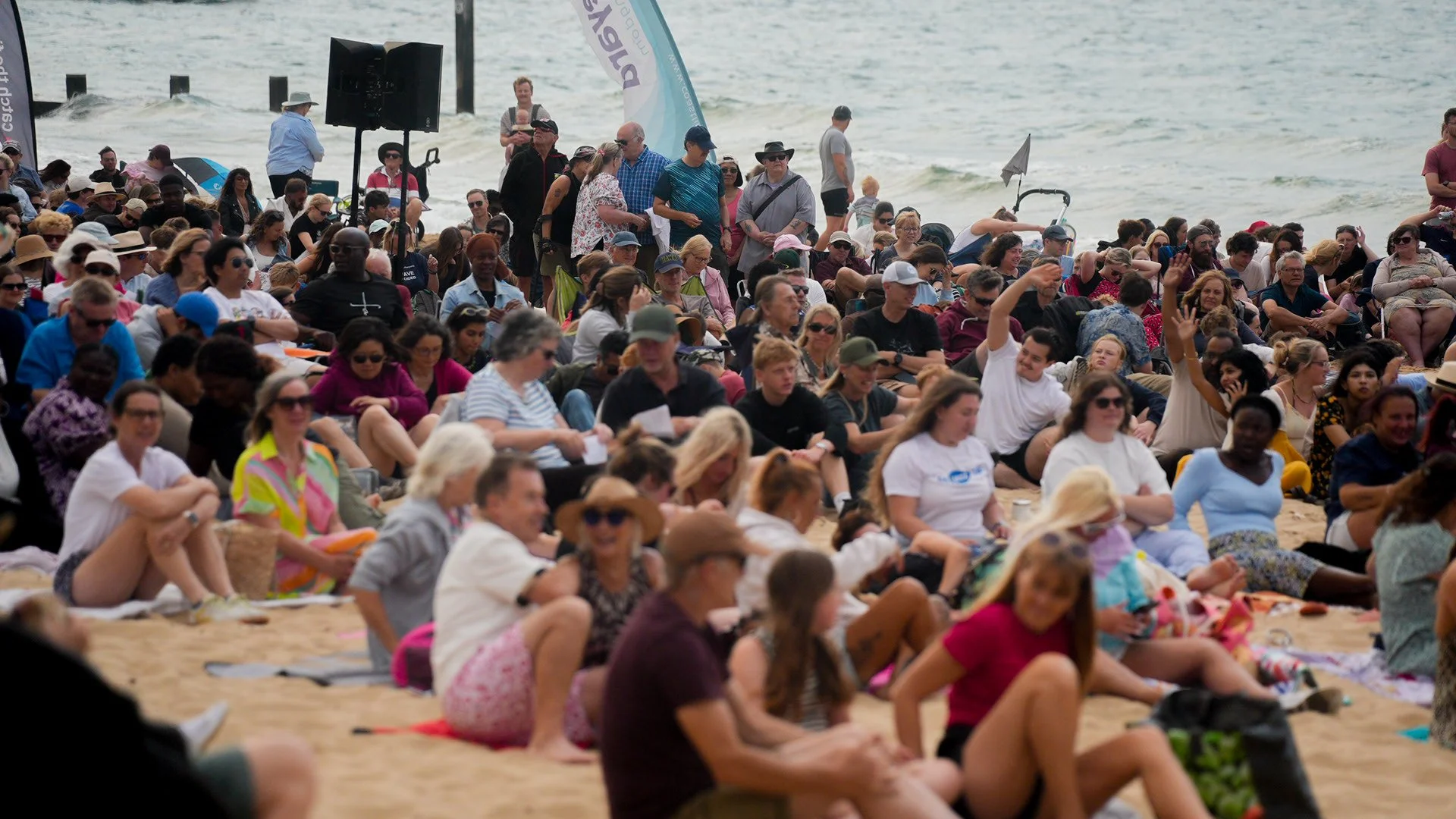 A large crowd of people gathered on a sandy beach near the ocean, sitting on blankets and chairs, with some standing and talking, while the ocean waves are visible in the background.